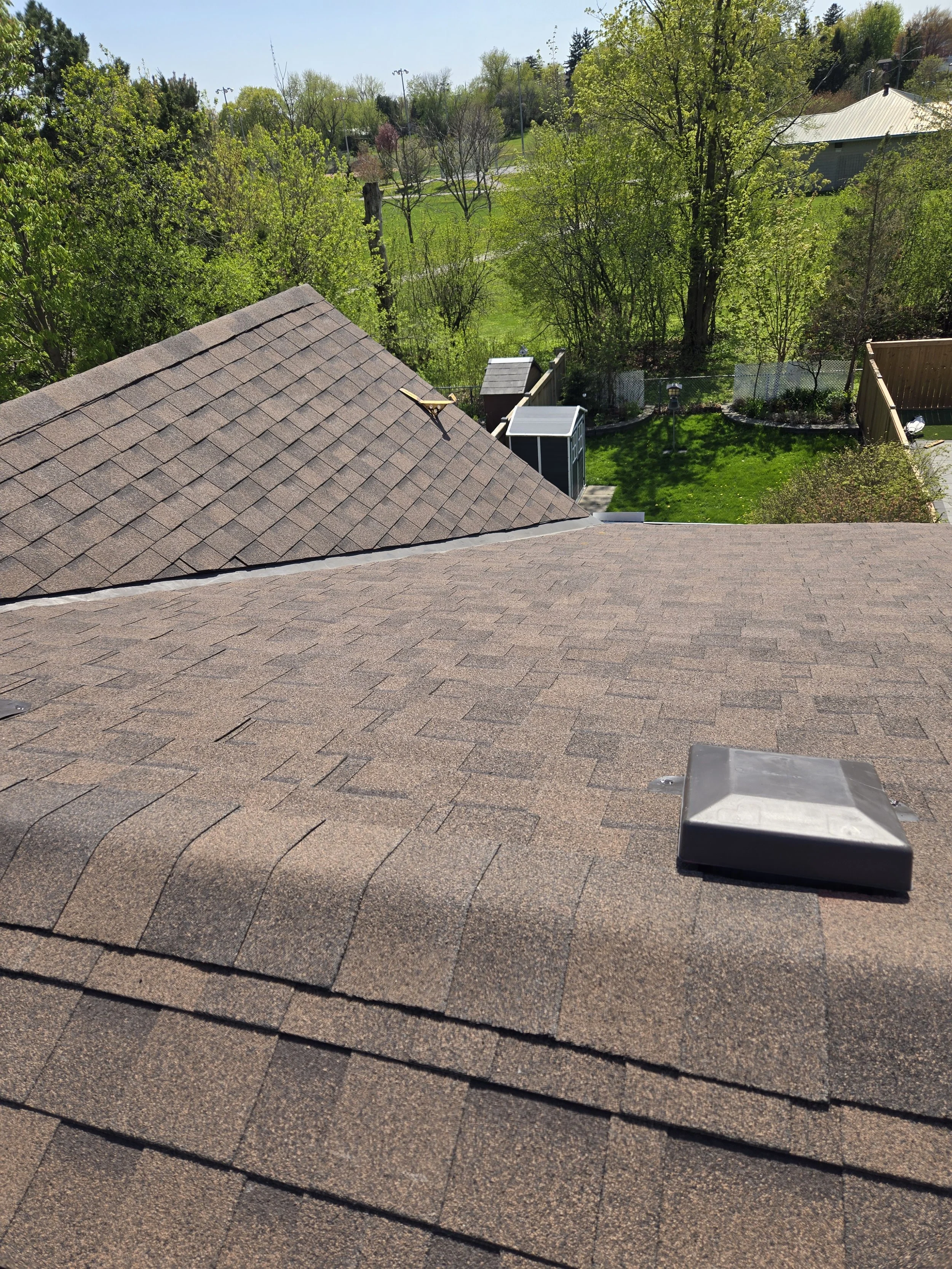 View of a residential roof with asphalt shingles, a chimney cap, and a roof vent, overlooking a backyard with green grass, trees, a small shed, and a garden area.