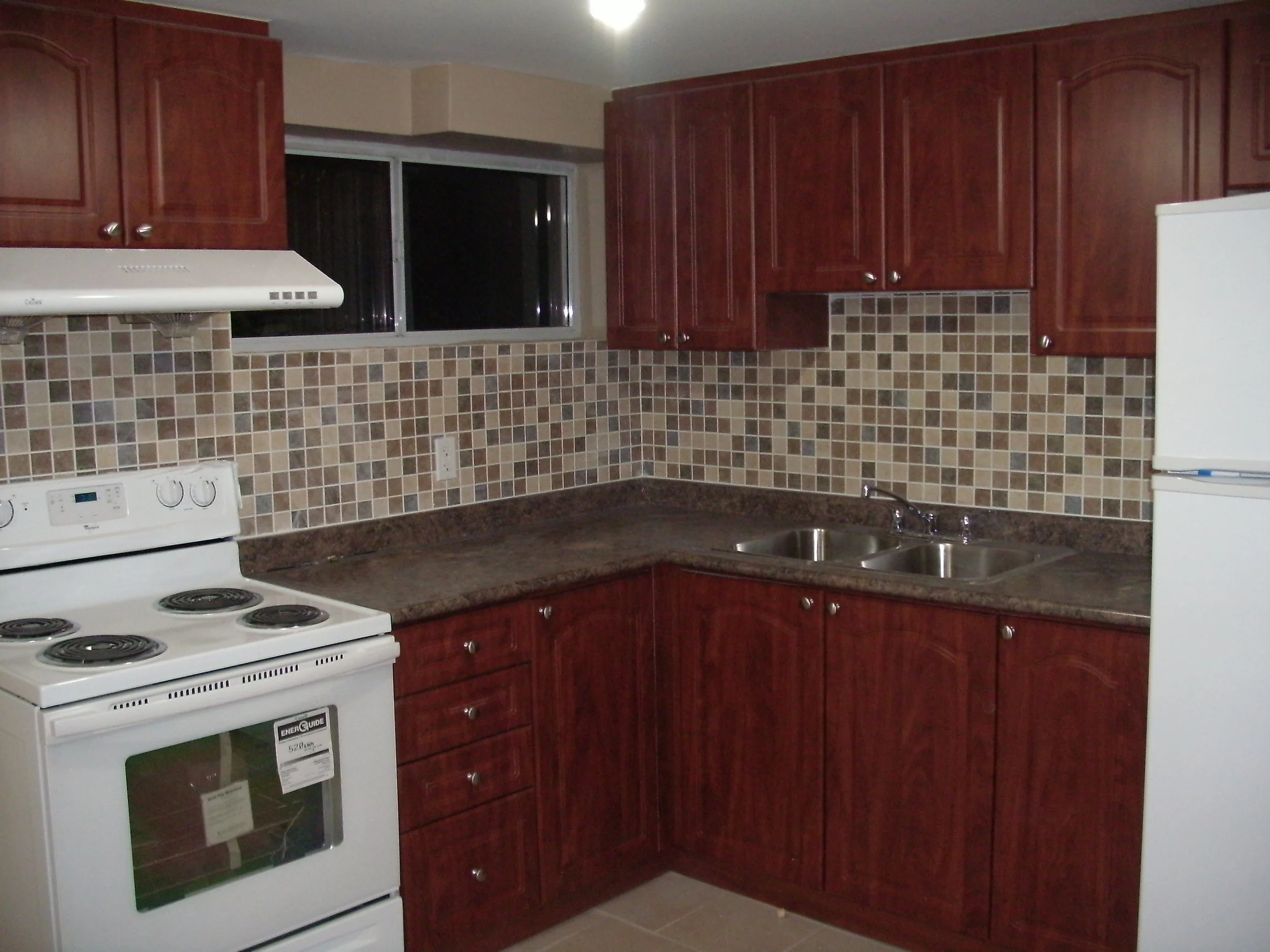 Kitchen with dark wood cabinets, a white stove on left and fridge on right, a greyish brown marbled counter, a tile backsplash of multiple brown shades, a sink, and a window.
