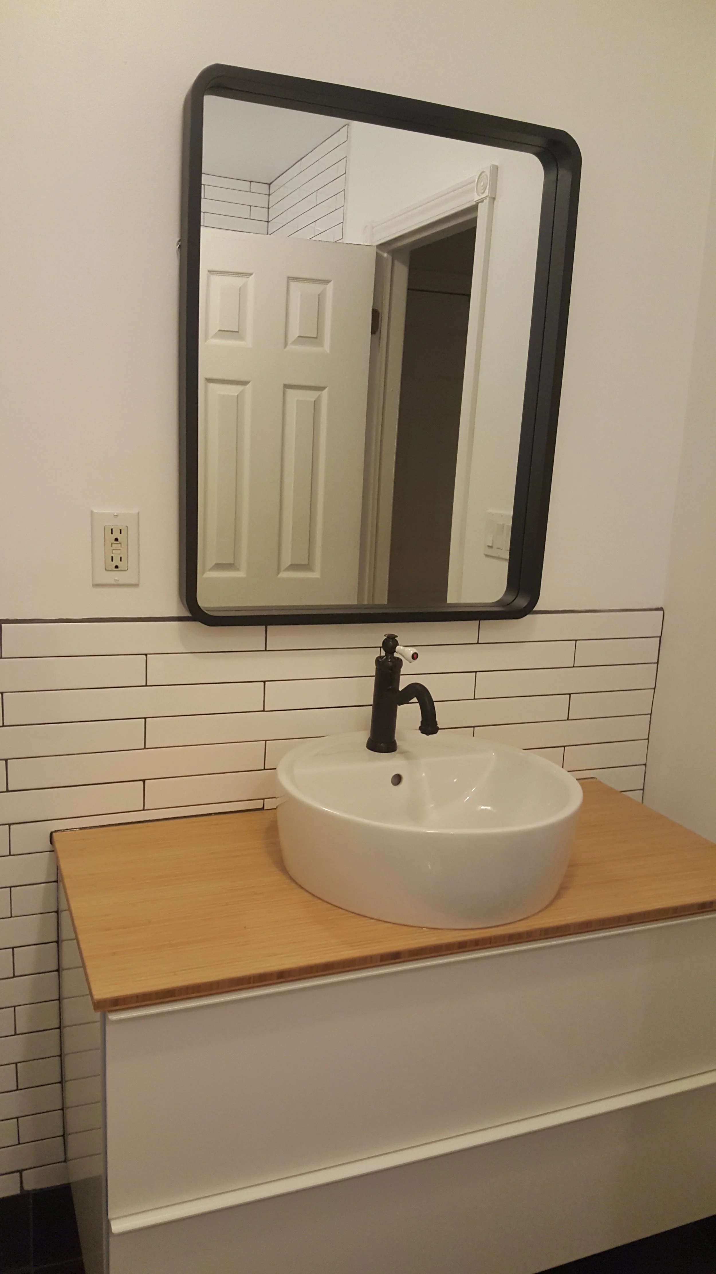 A bathroom with a round white sink and a black faucet on a light brown wooden countertop, a mirror above the sink with a black border, white tile backsplash, and a white door reflected back in the mirror.