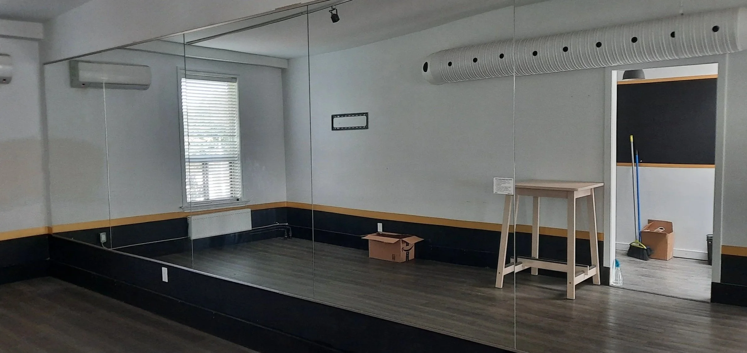 An empty mirror yoga studio with a wooden floor, white walls, a window with blinds, and a cardboard box with some cleaning supplies and a mop leaning against the wall.