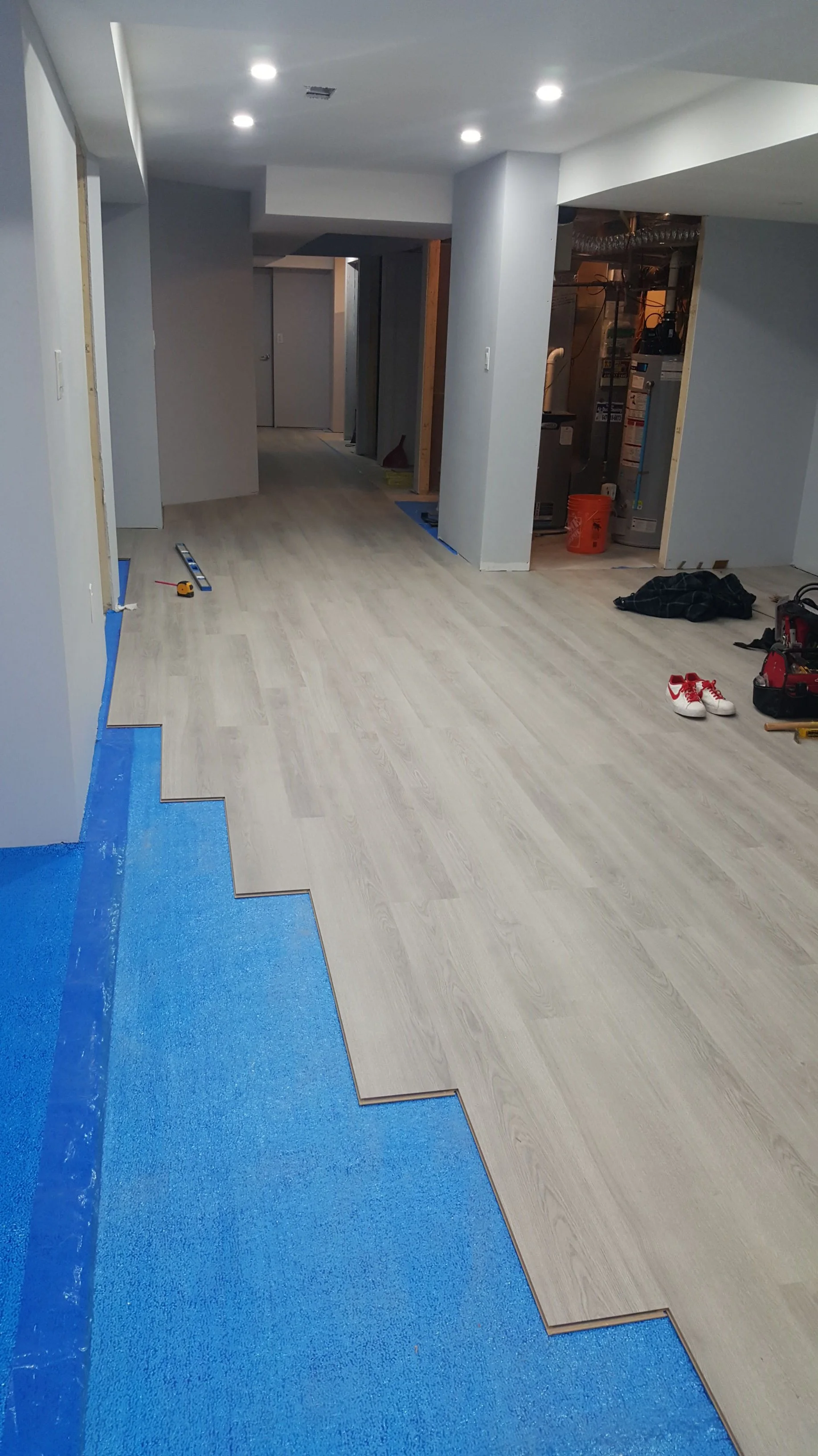 Renovation with new flooring being installed. Blue painter's tape is on the floor below the new light-colored wood laminate flooring. Nearby tools and construction materials are visible.