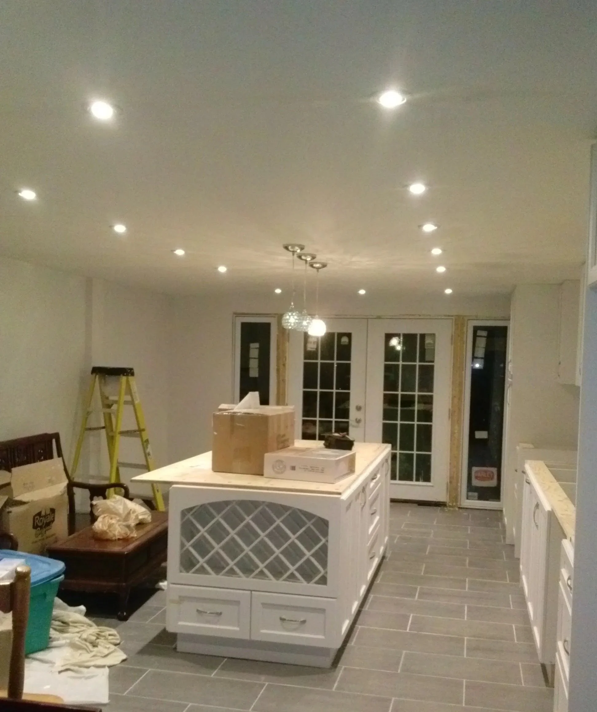 Kitchen with a white island, a ceiling with multiple pot lights, a hanging light fixture, sliding glass door, and various boxes and items scattered around.