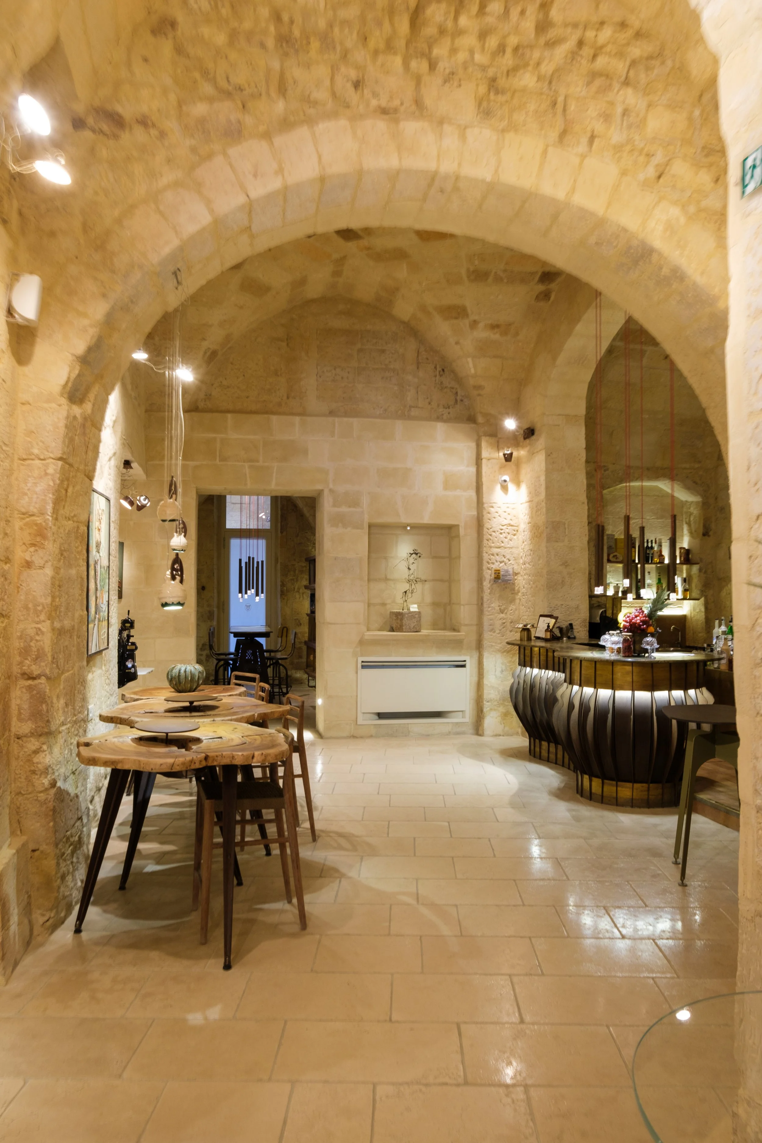 A cozy restaurant or coffee shop interior with stone walls and arched ceilings. It features a wooden table with four chairs, and a bar area with bottles and flowers in a vase. Warm lighting highlights the stone architecture.