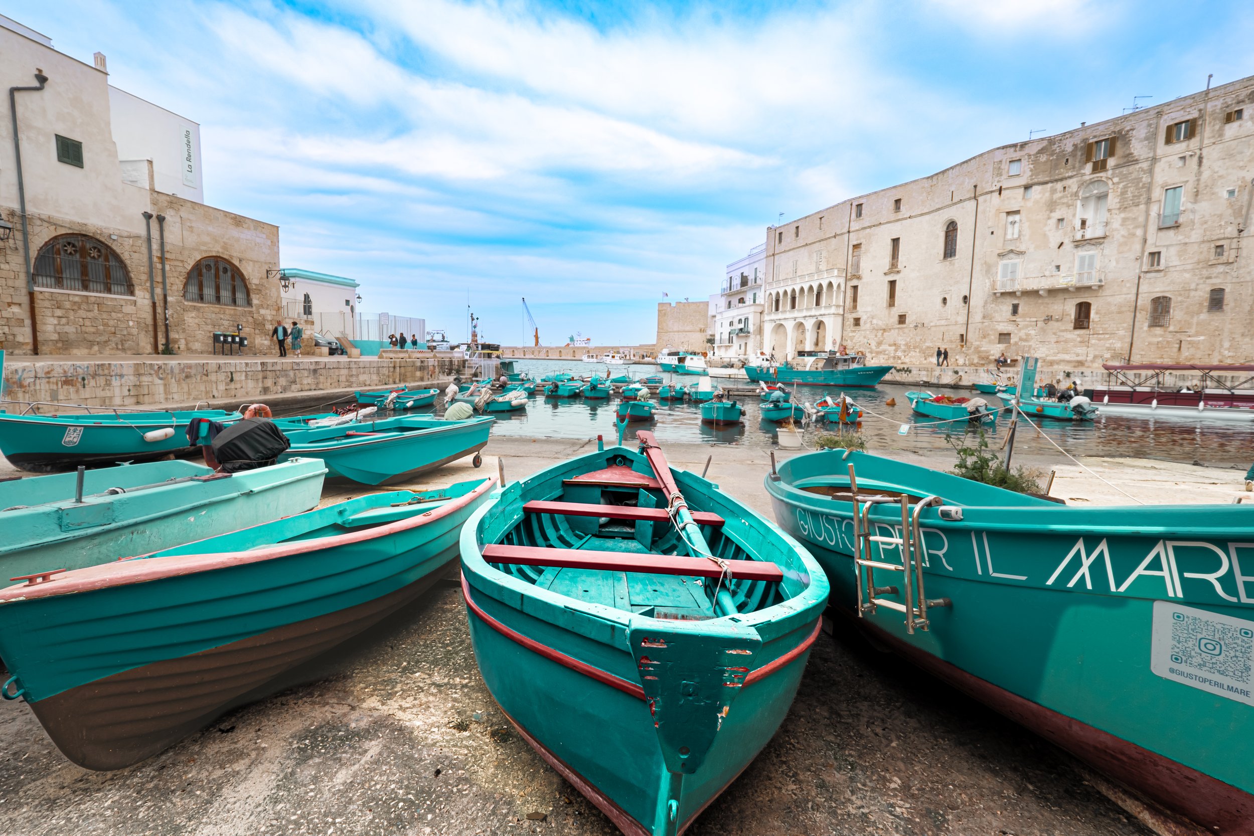 Several small turquoise boats docked along a harbor with historic stone buildings and a cloudy sky in the background.