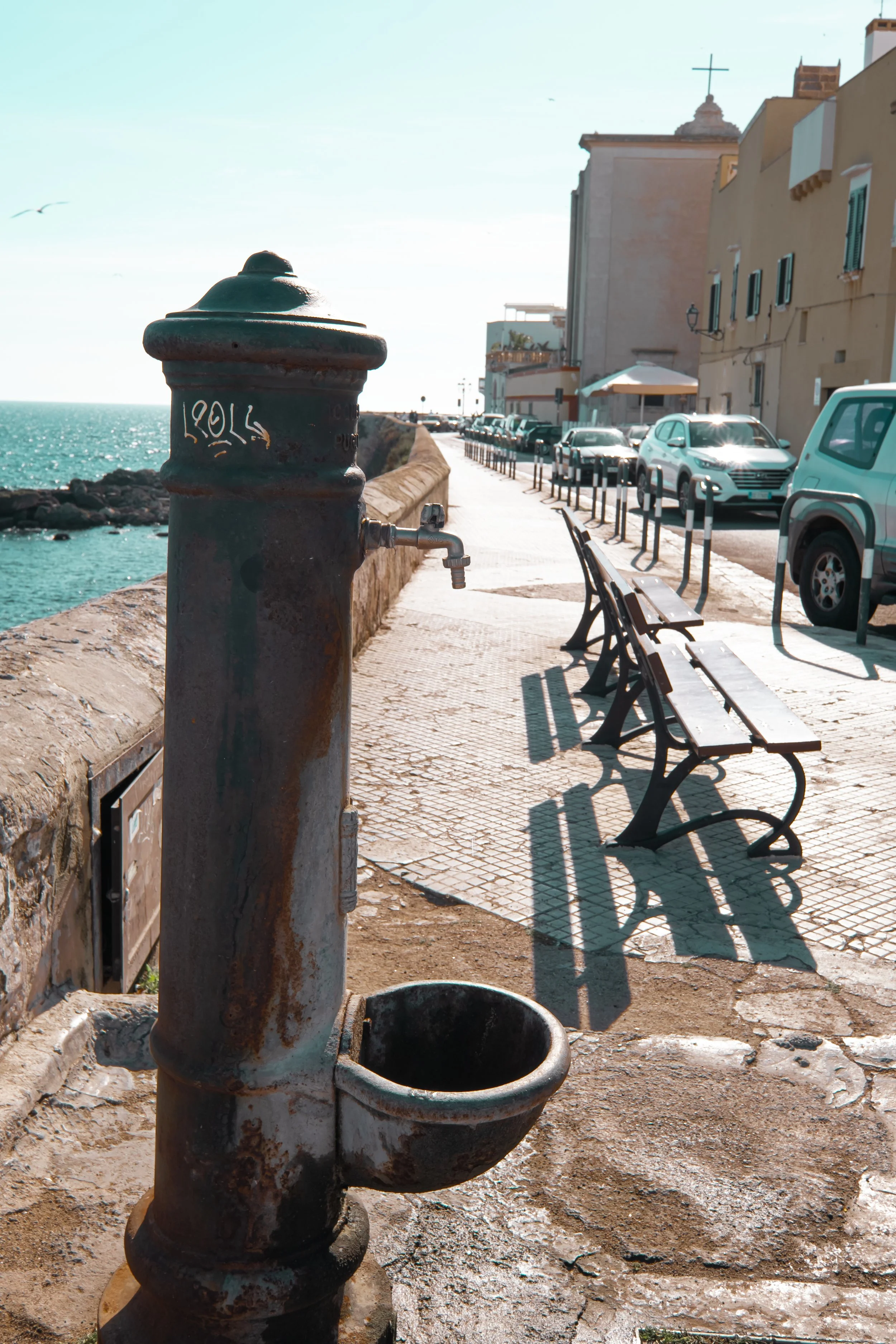 Old pipe with a faucet on a seaside promenade, parked cars, benches, and buildings with a cross on top in the background.