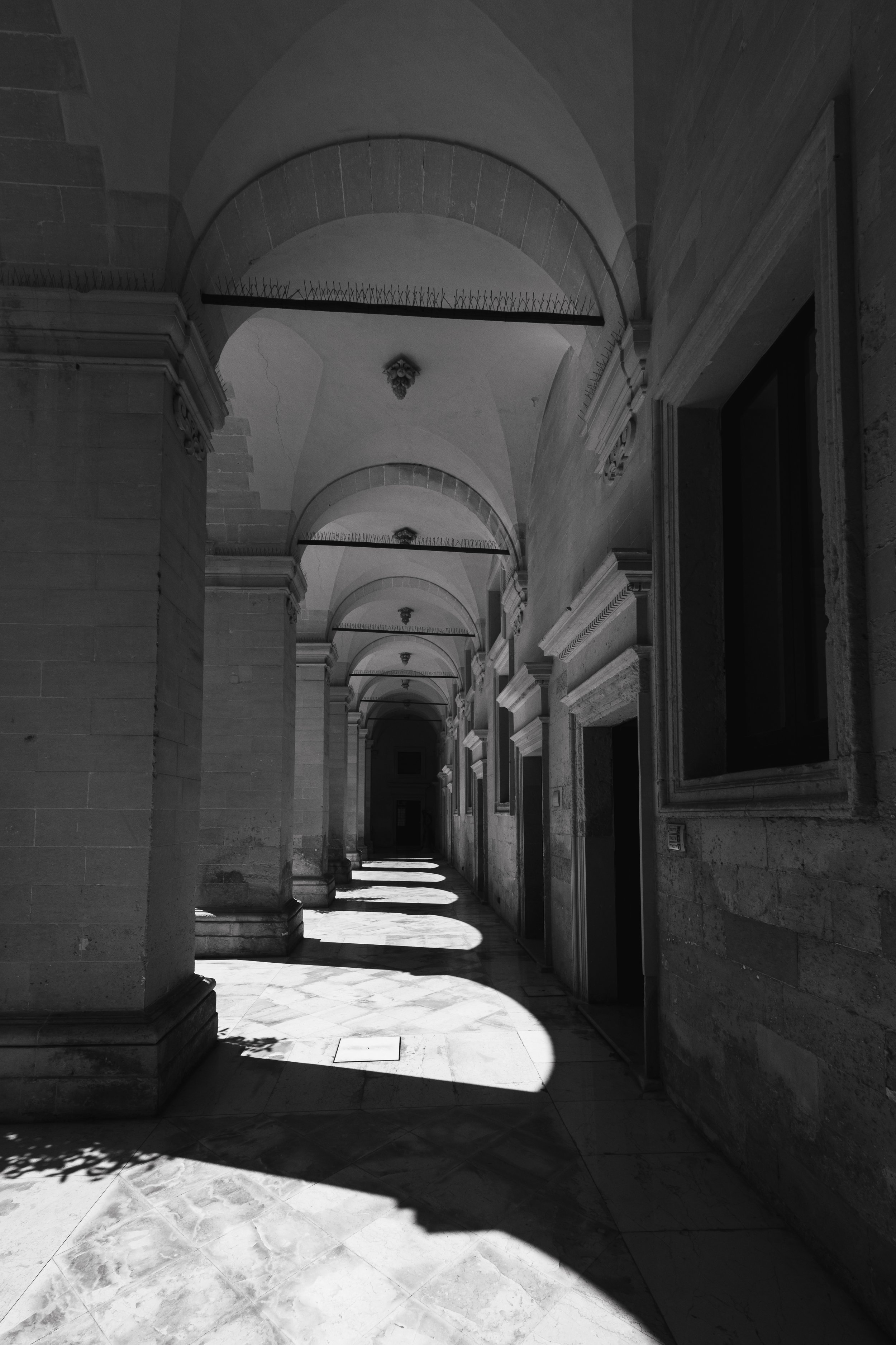 A long corridor with arched ceilings and stone walls, sunlight casting shadows on the marble floor, and ornate light fixtures hanging from the ceiling.