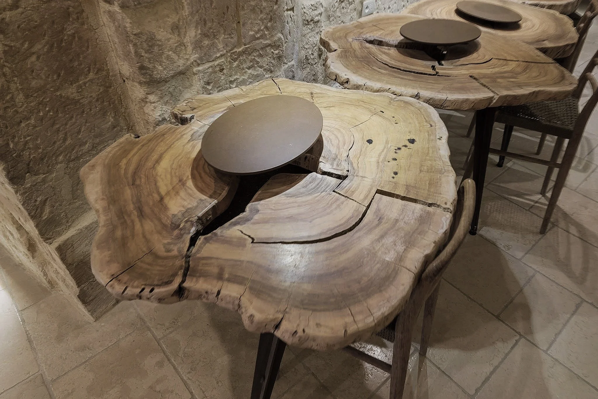 A rustic dining table made from a large, natural wood slab with a live edge, paired with black chairs, set against a stone wall.