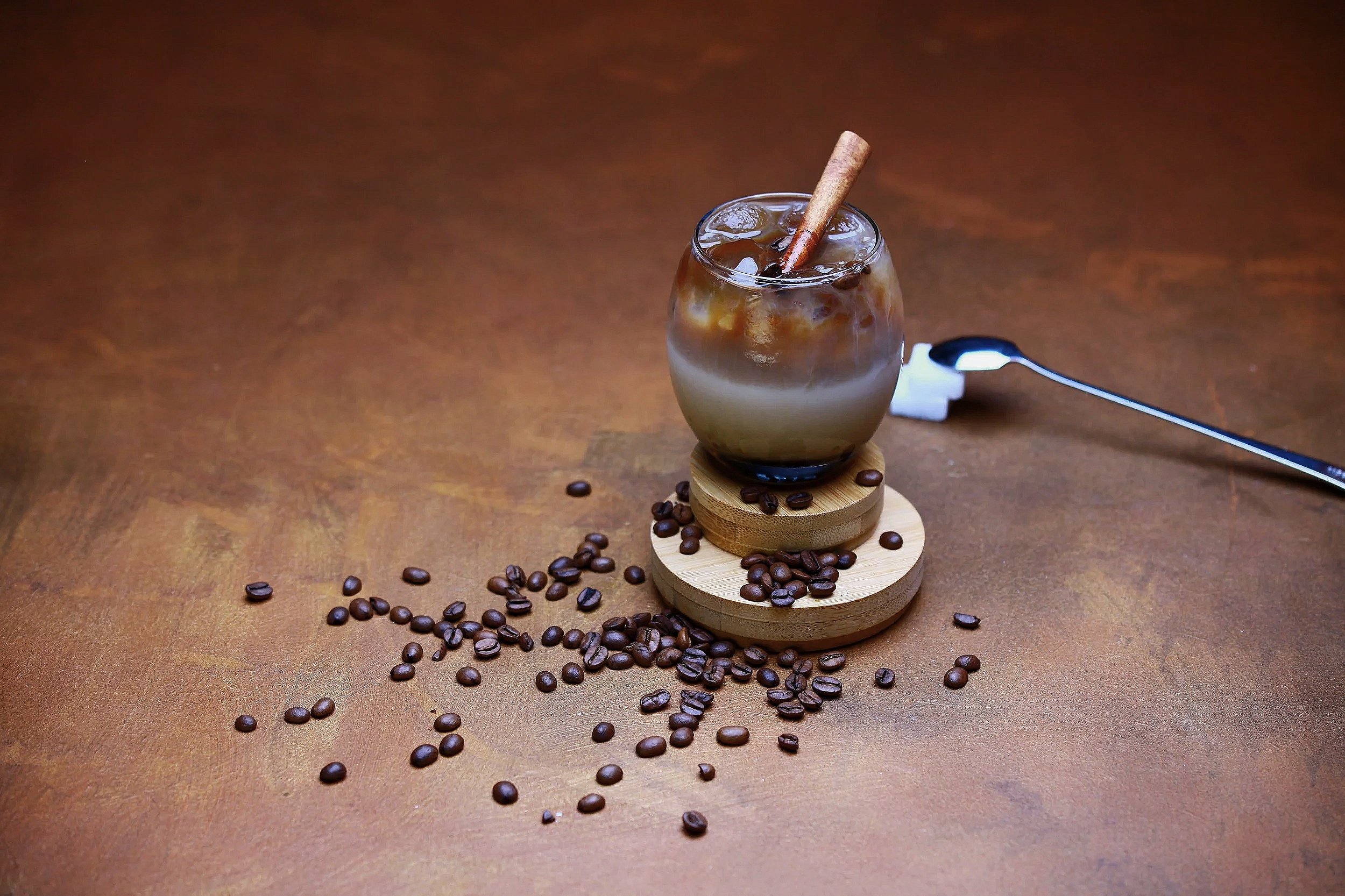 A glass of iced coffee with ice cubes and a cinnamon stick, surrounded by scattered coffee beans on a wooden surface.