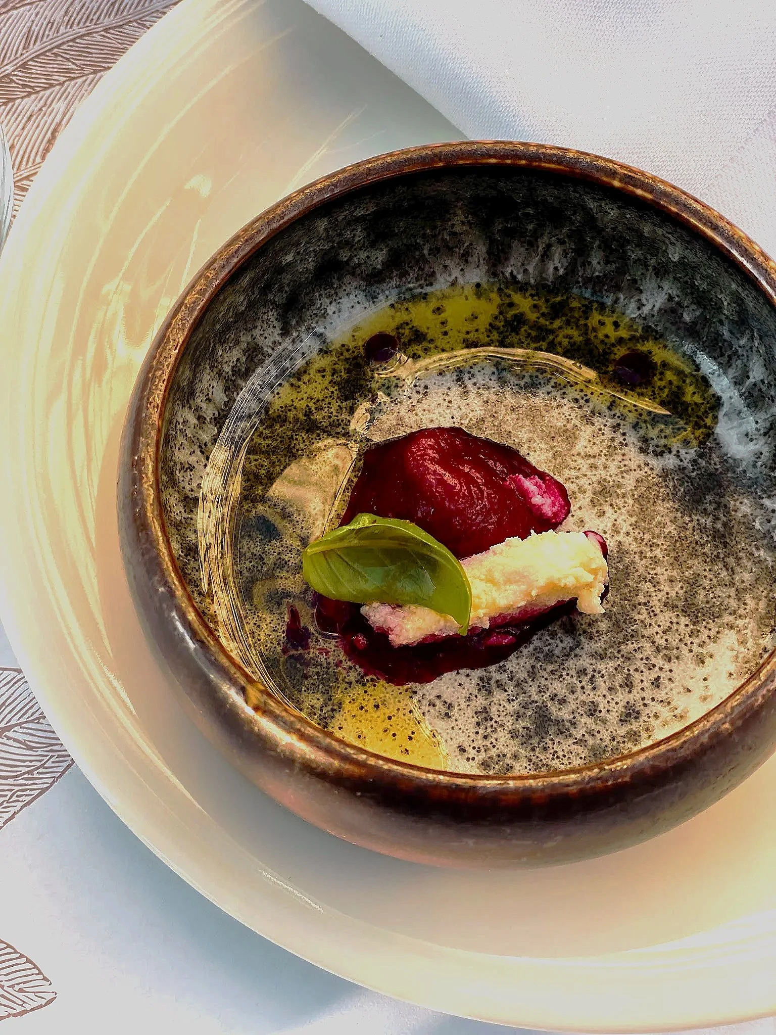 Close-up of a ceramic bowl with a soot-stained interior holding a mixture of dark oil and a red substance topped with a small green leaf, placed on a white plate on a table with a white and textured placemat.