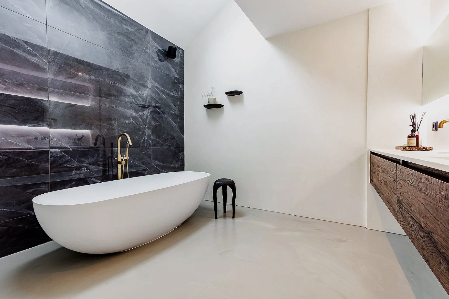 Modern bathroom with free-standing bathtub, black stool, dark gray marble accent wall, white walls, minimalist black shelves, wooden vanity with soap dispenser
