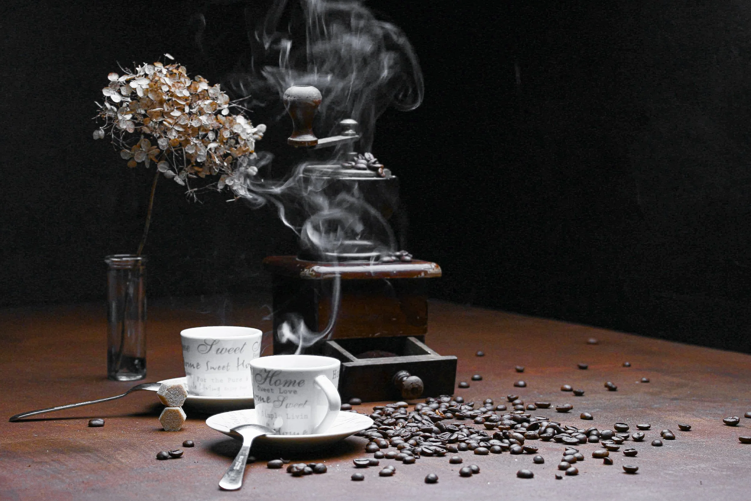 A wooden table with scattered coffee beans, a spoon, and two white cups with coffee-related words, with a vintage coffee grinder, a dried hydrangea flower in a glass, and steam rising from the grinder in a dark background.