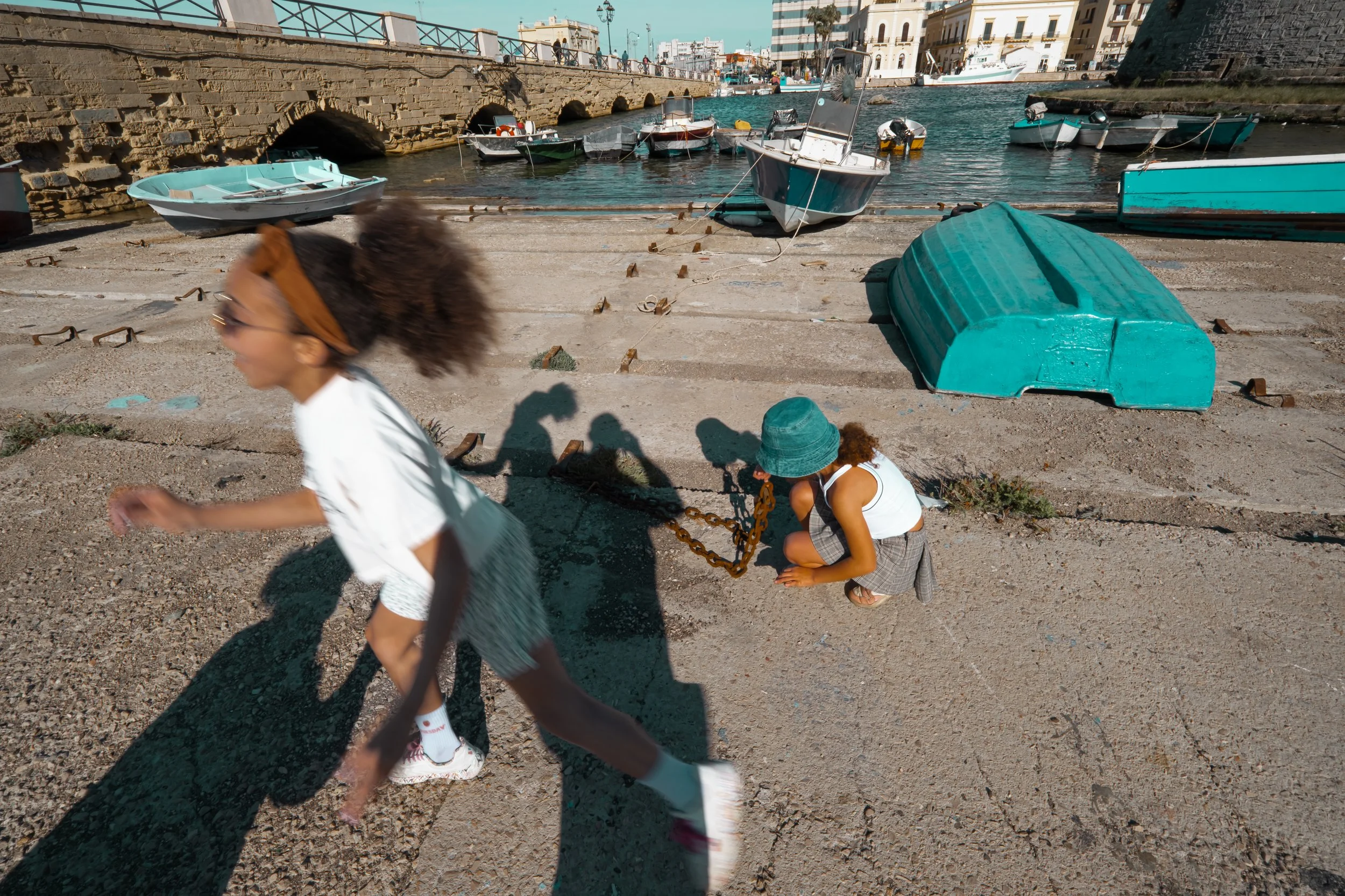 Two children sitting and playing near the water with boats docked in the background at a scenic harbor or marina.