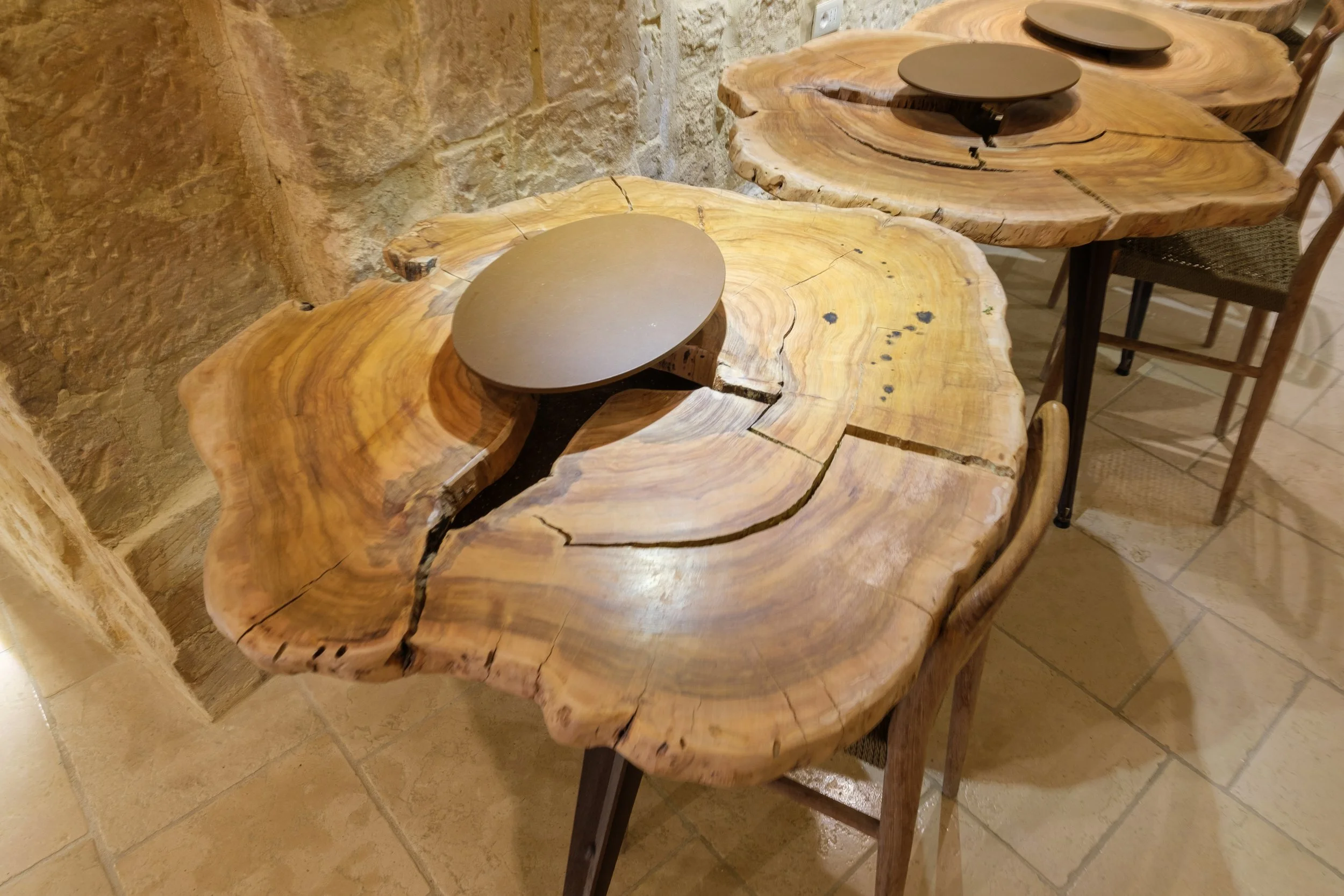 Set of three rustic wooden tables with irregular edges and visible natural wood grain, each with a round, dark-colored plate on top, arranged against a stone wall and tiled floor.