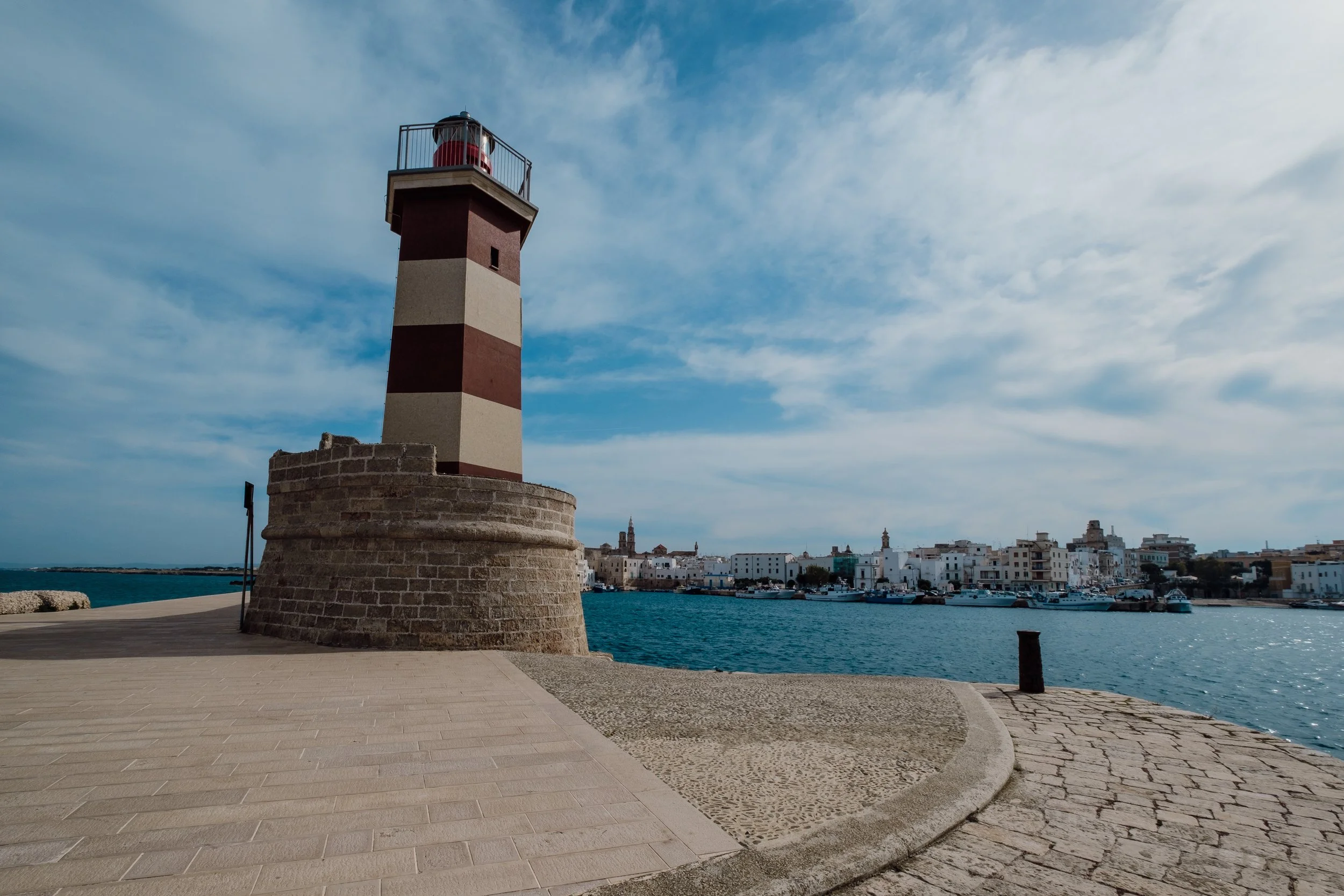 A lighthouse with red and white stripes on a stone base, overlooking a harbor with white buildings and boats, under a partly cloudy sky.