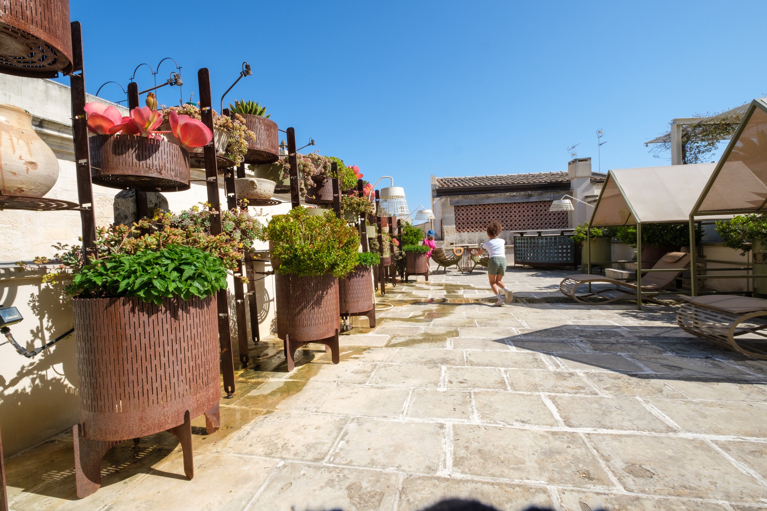Outdoor terrace with potted plants on a wall-mounted metal rack, two children playing, lounge chairs, and a clear blue sky.