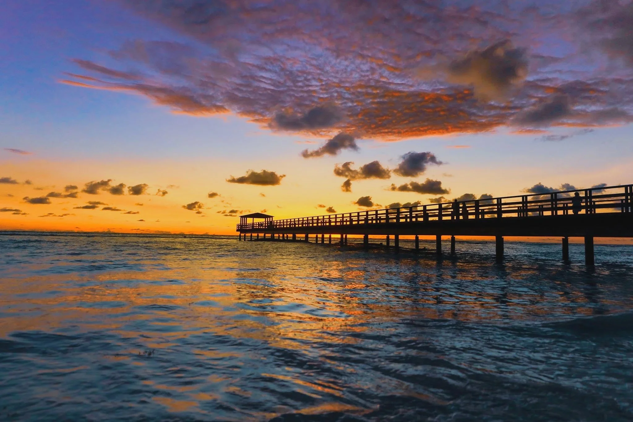 Sunset over the ocean with a wooden pier extending into the water and a small gazebo at the end. The sky is painted with shades of orange, purple, and blue, and there are scattered clouds.