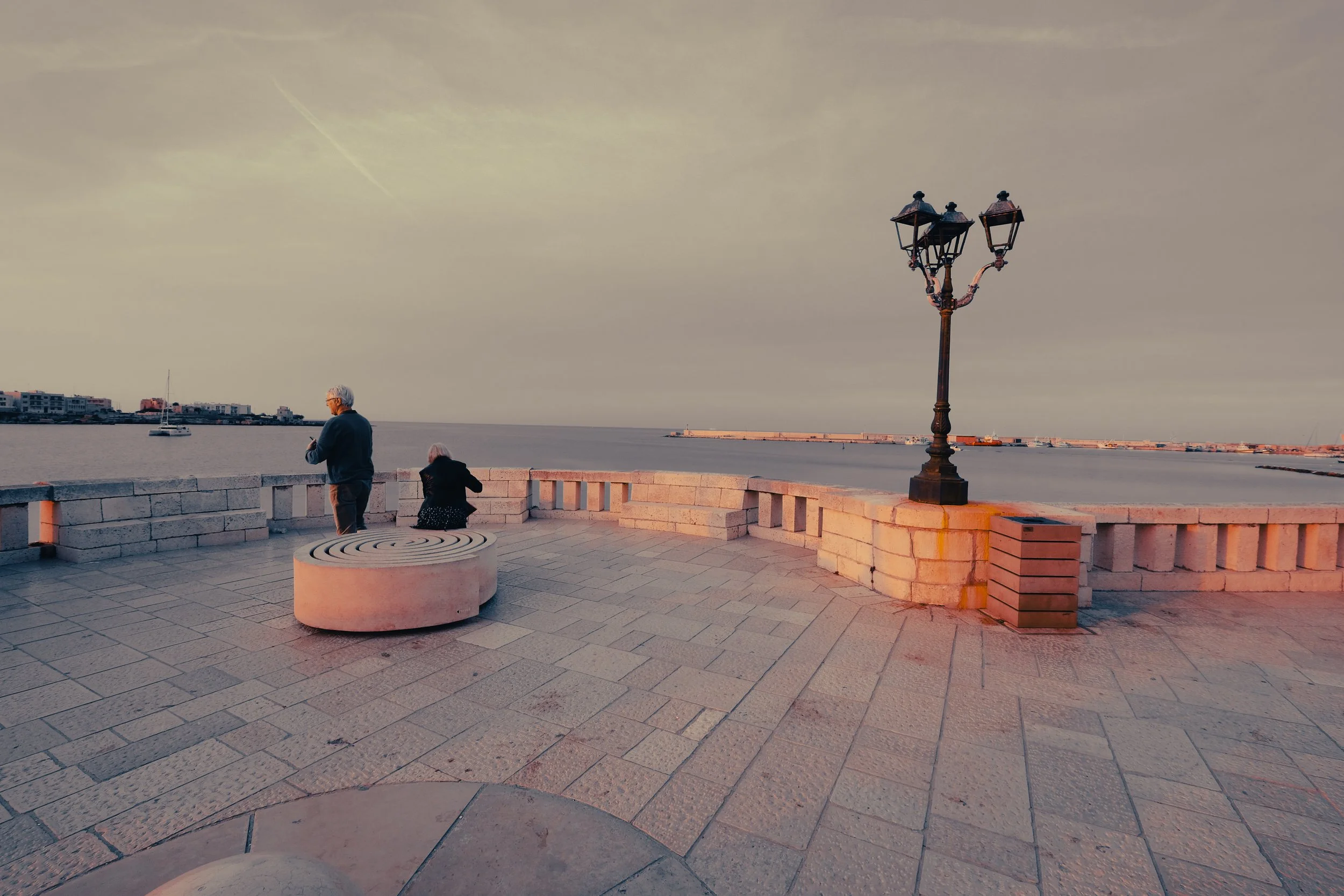 People standing near a stone balustrade overlooking a body of water with a sailboat and a distant shoreline under a cloudy sky.
