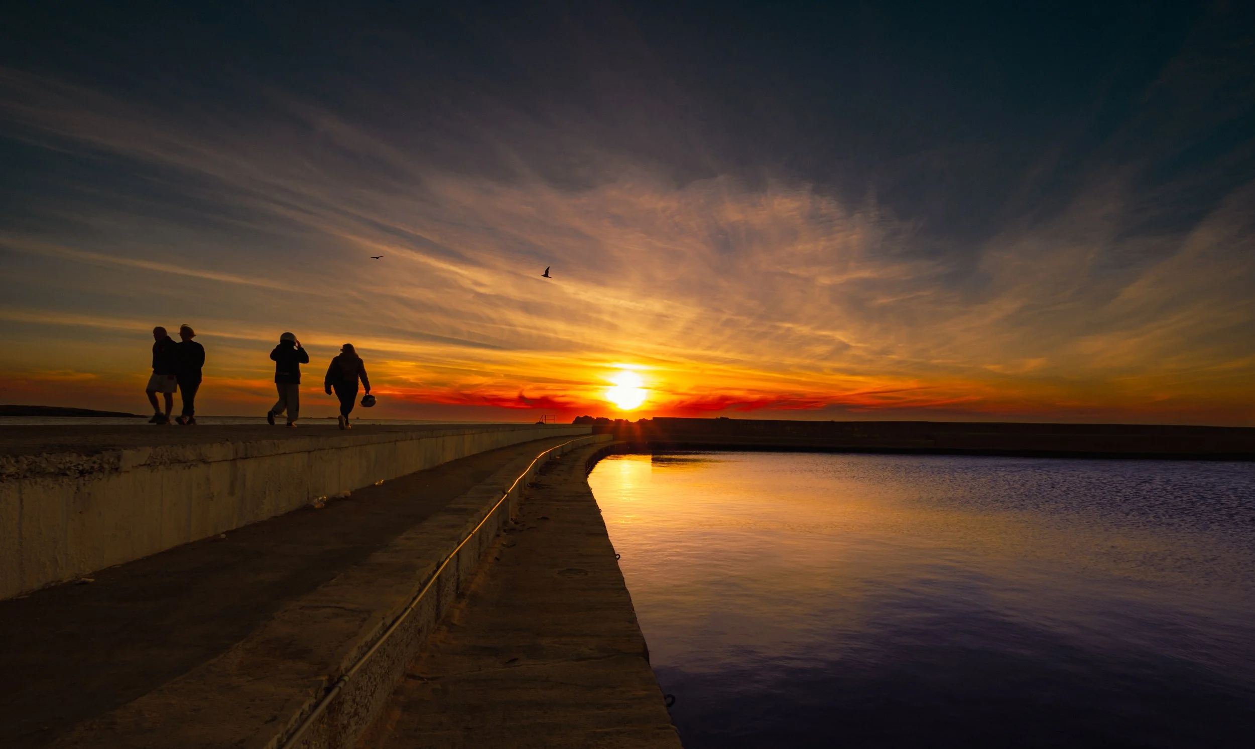 Silhouettes of four people walking along a concrete pier at sunset, with a colorful sky and birds flying overhead.