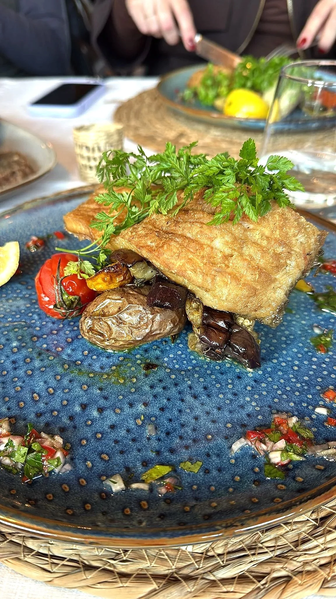A plated dish of crispy fried fish topped with fresh parsley, served with roasted cherry tomatoes and eggplant, and garnished with lemon slices, on a blue textured plate.
