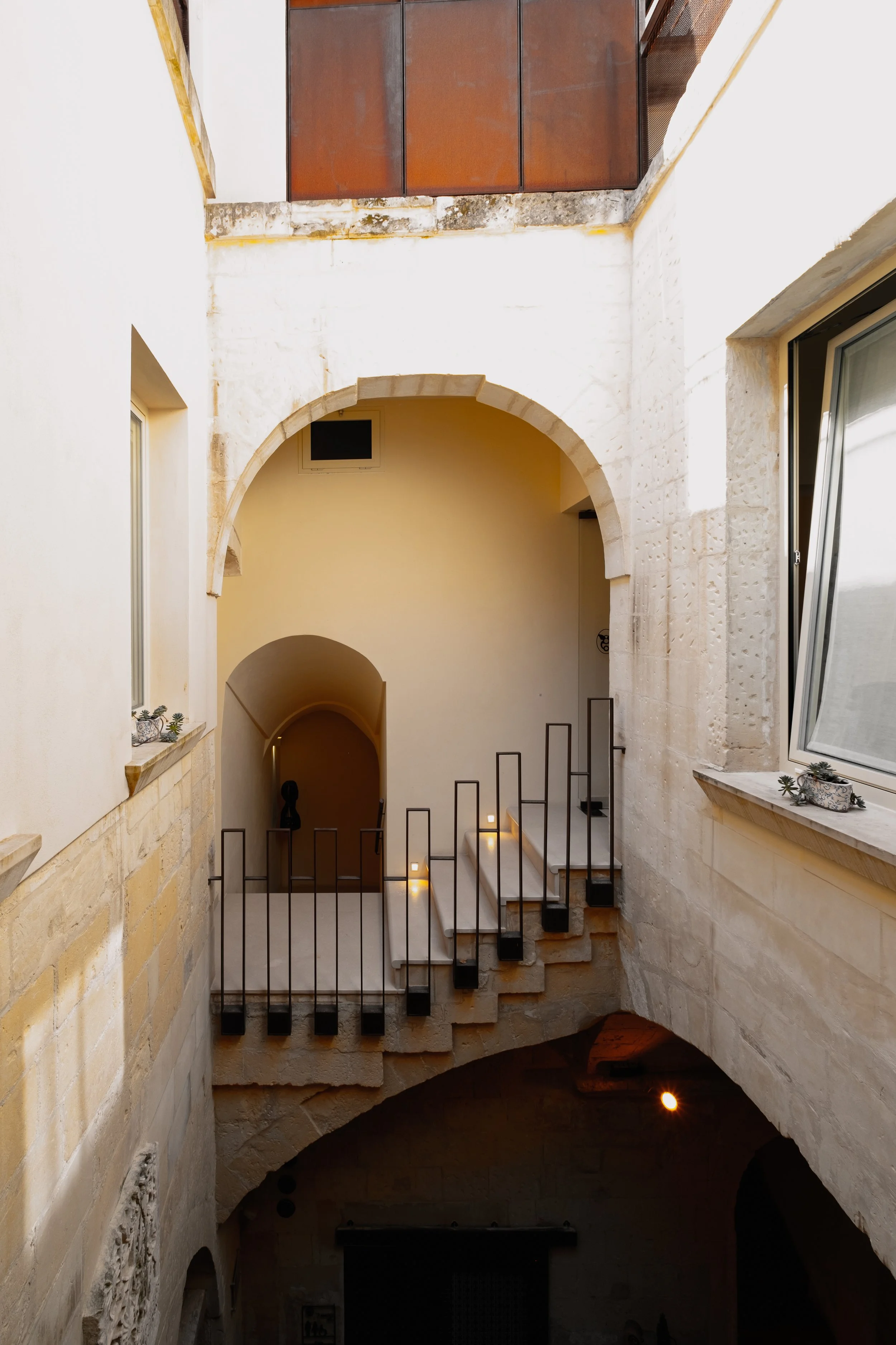 An indoor historical or architectural site with stone walls, an archway, stairs, and interior balcony with a metal railing. A doorway and small window are visible, with minimal decorative elements and modern lighting illuminating the area.
