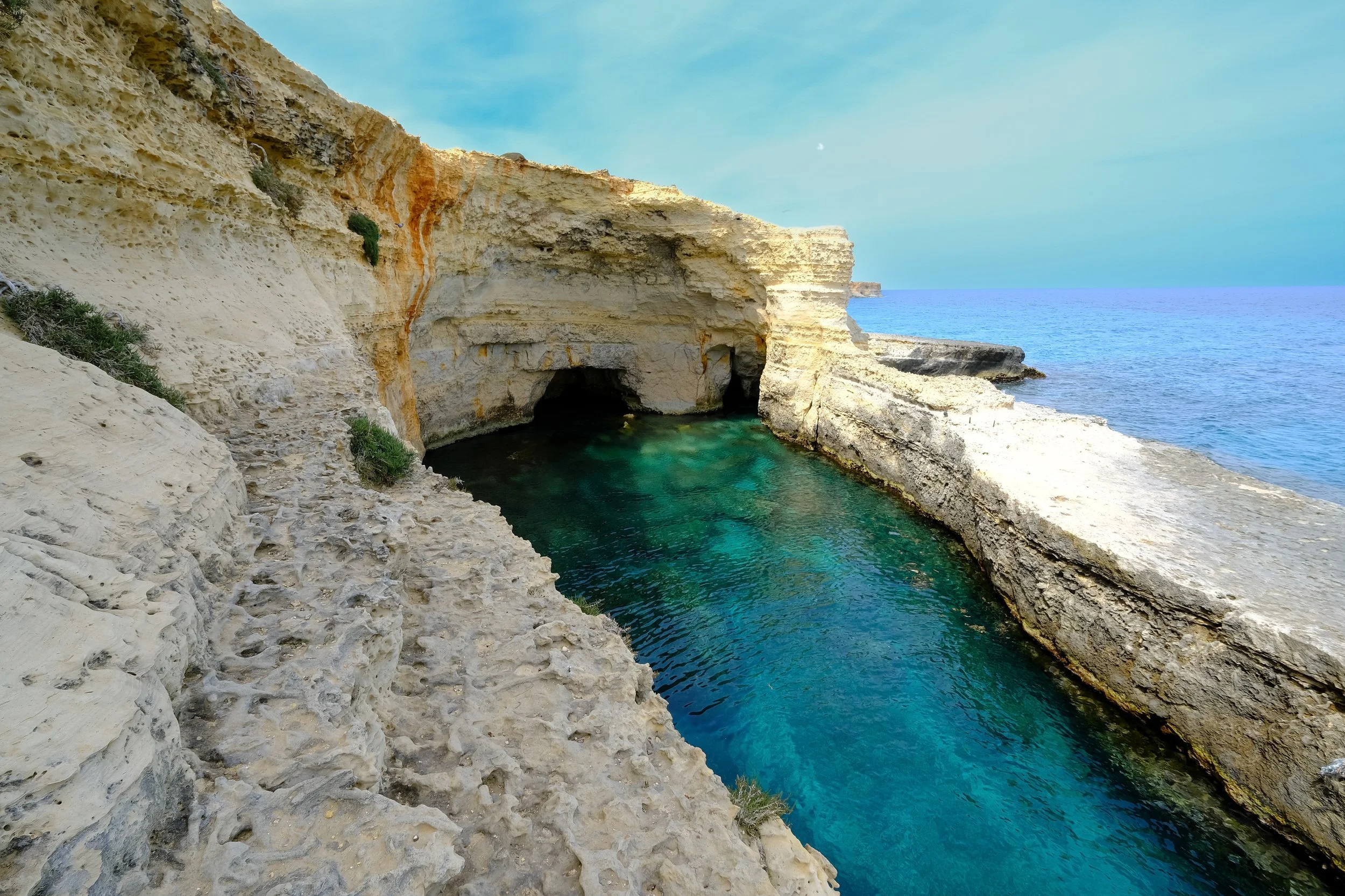 Cliffs with a cave opening and turquoise water at the base, overlooking the ocean under a partly cloudy sky.