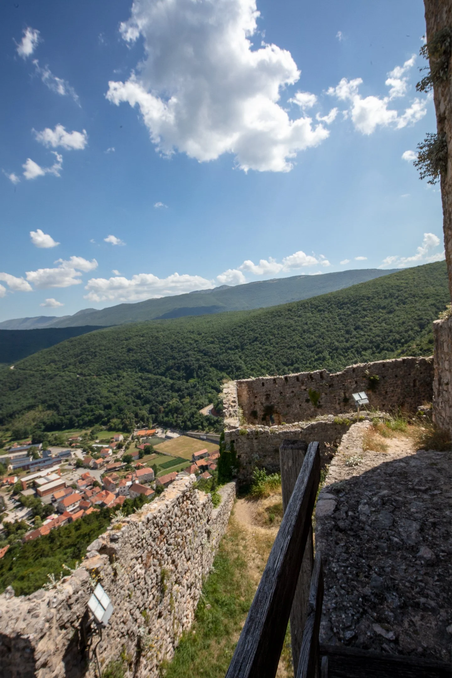 Scenic landscape with green hills, a small town with red-tiled roofs, a stone fortress wall in the foreground, and a partly cloudy blue sky.