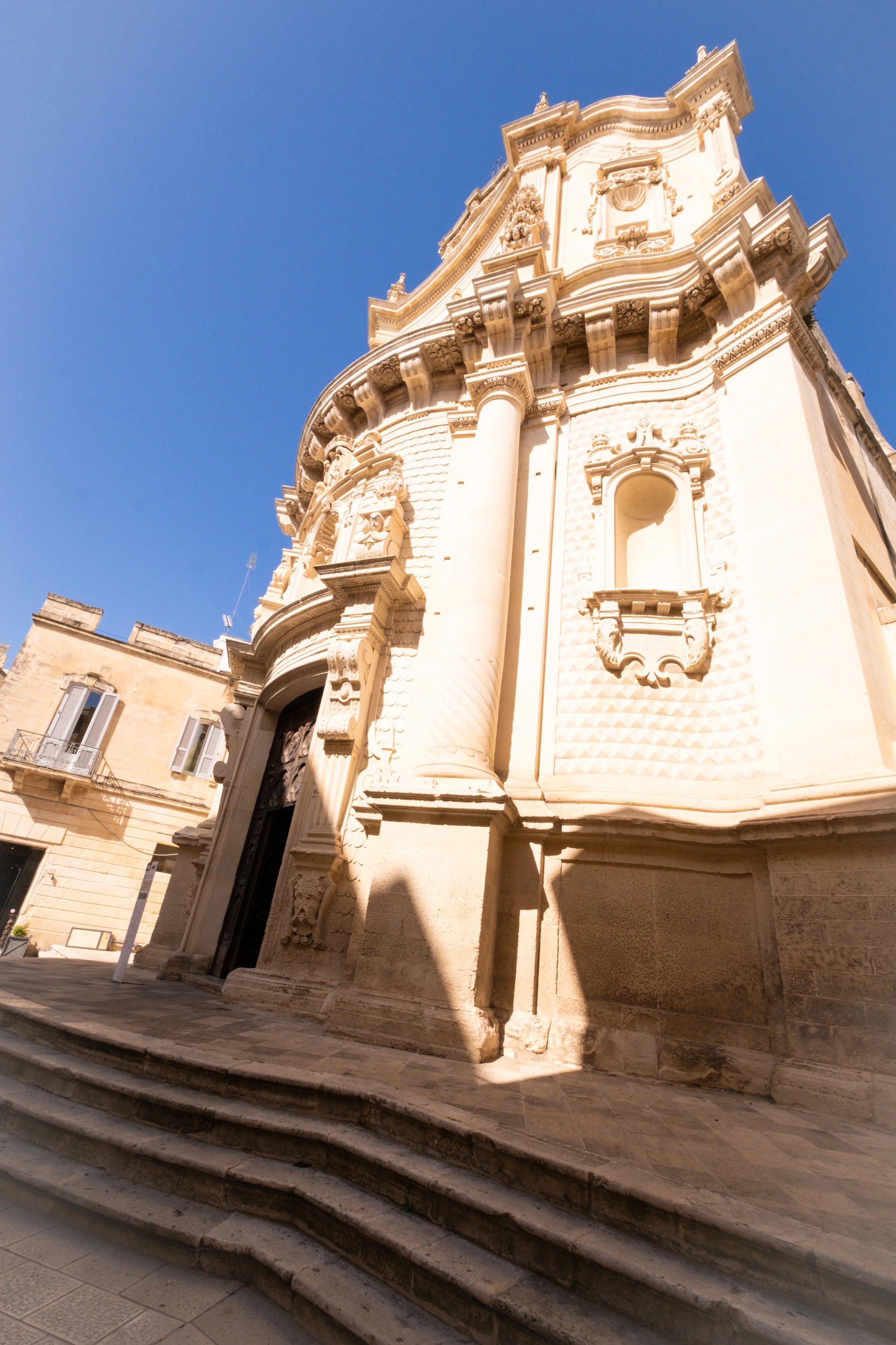 Exterior of a historic baroque church with ornate architectural details, including columns, decorative busts, and a curved facade, set against a clear blue sky.