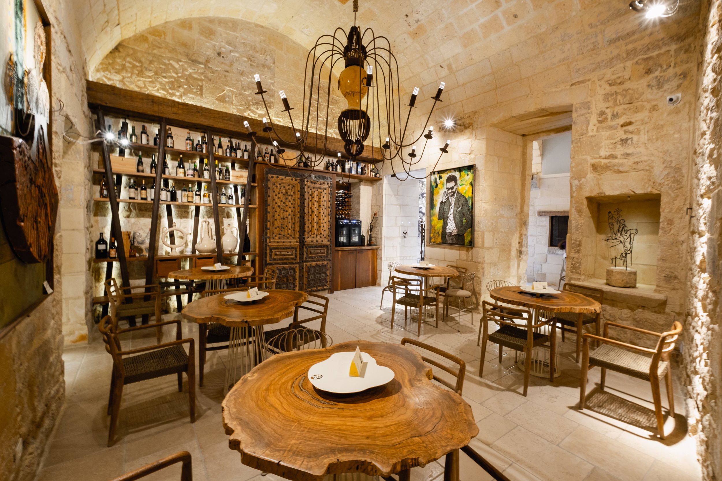Interior of a rustic restaurant or cafe with wooden tables and chairs, stone walls, and decorative lighting, including a chandelier with exposed bulbs.