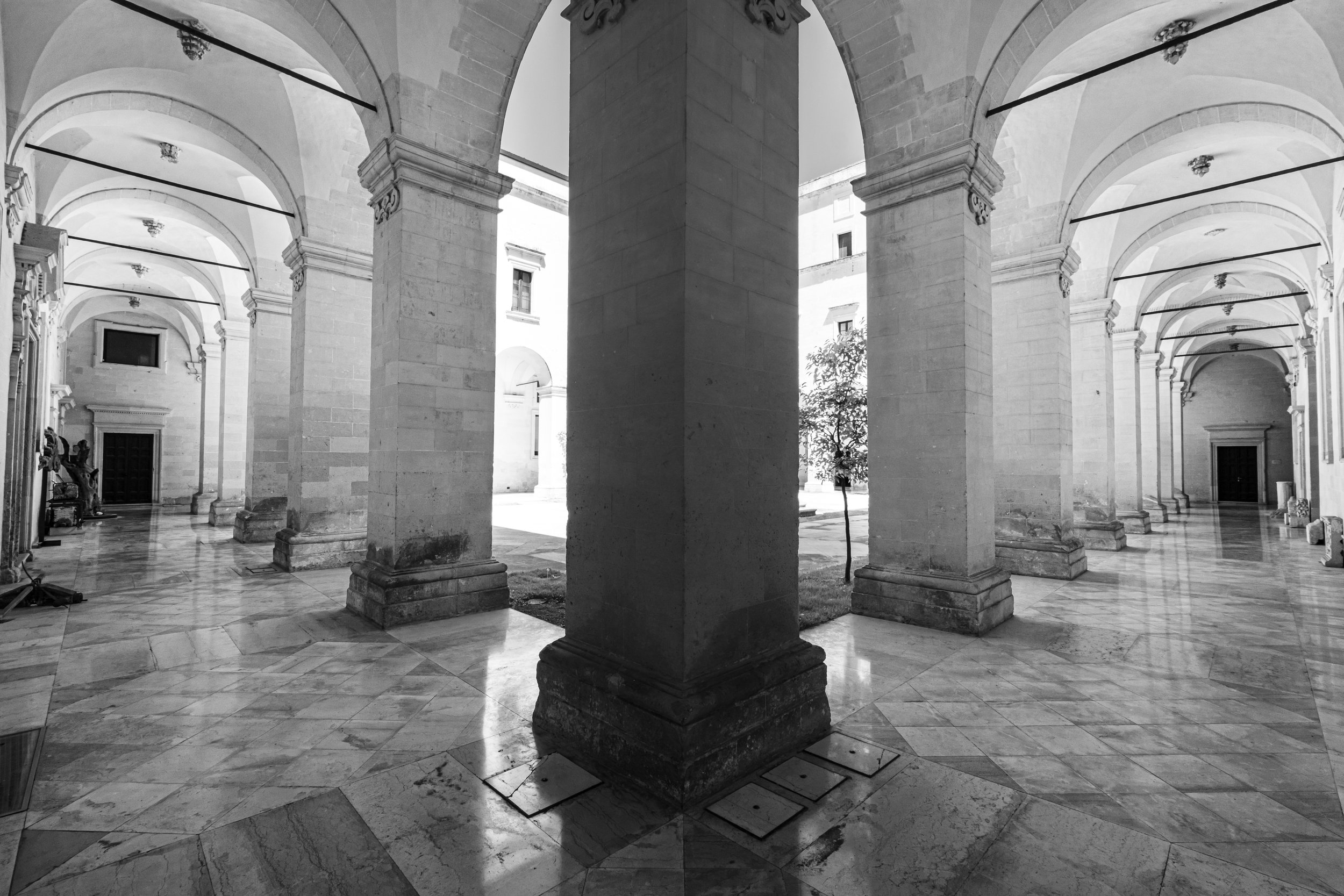 A black and white photograph of a classical architectural courtyard with large stone columns, arched ceilings, and a marble floor.