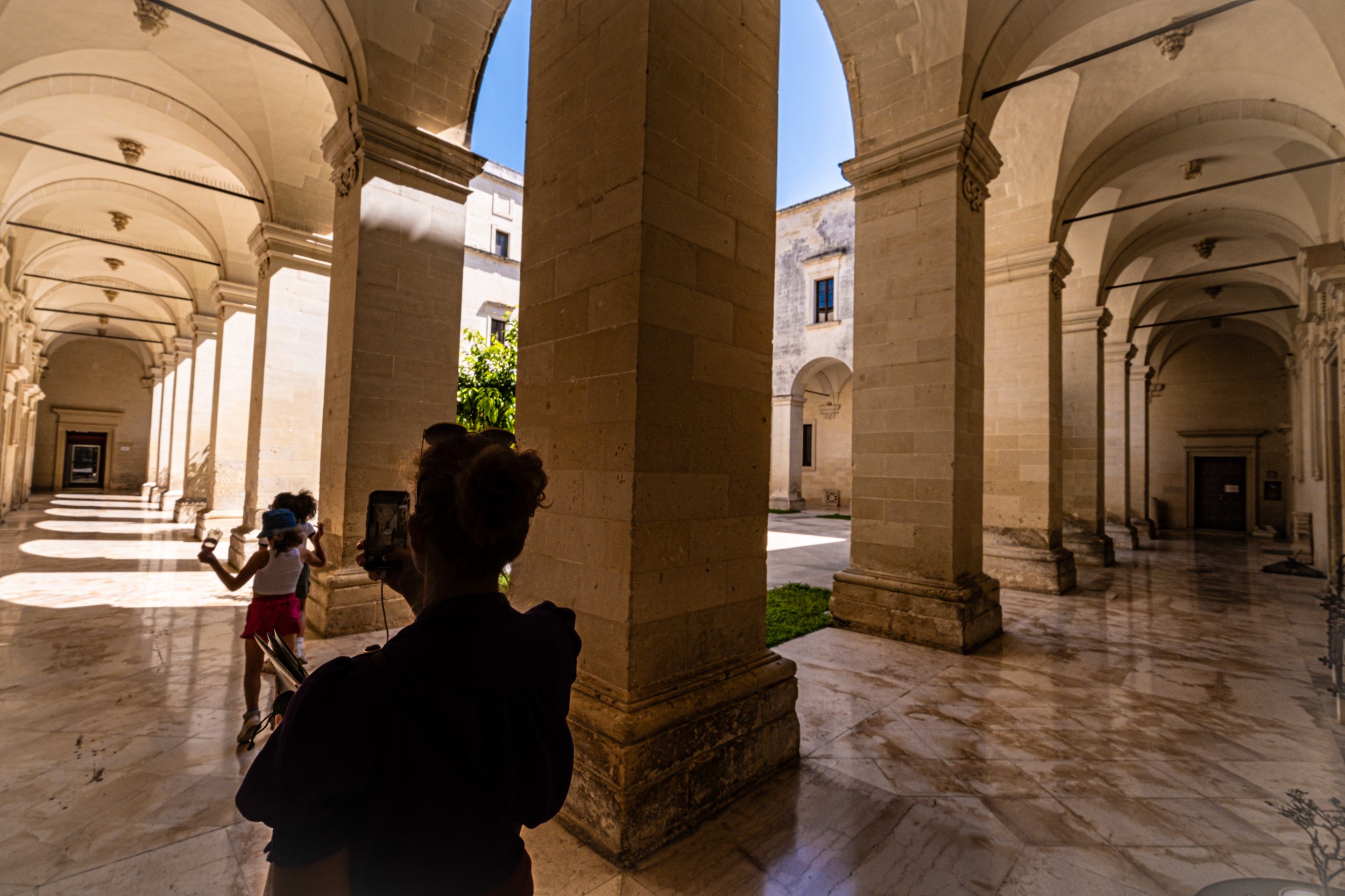 People inside a historic building with stone columns and arches, sunlight casting shadows, a child taking a photo, and a woman wearing a hat with another person in the foreground.
