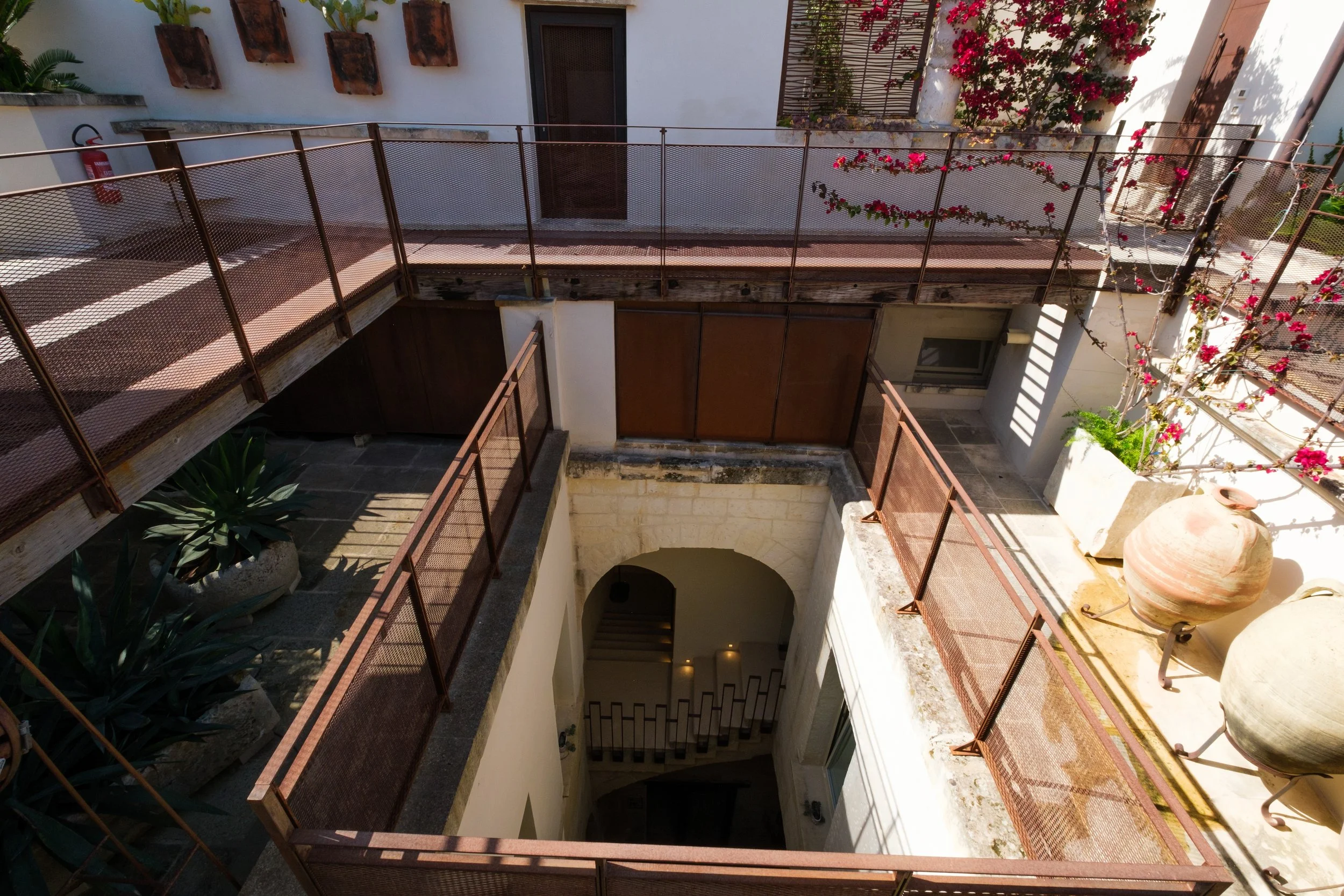 View of a Mediterranean-style courtyard with white walls, potted plants, bougainvillea climbing on the wall, and metal railings around the perimeter.