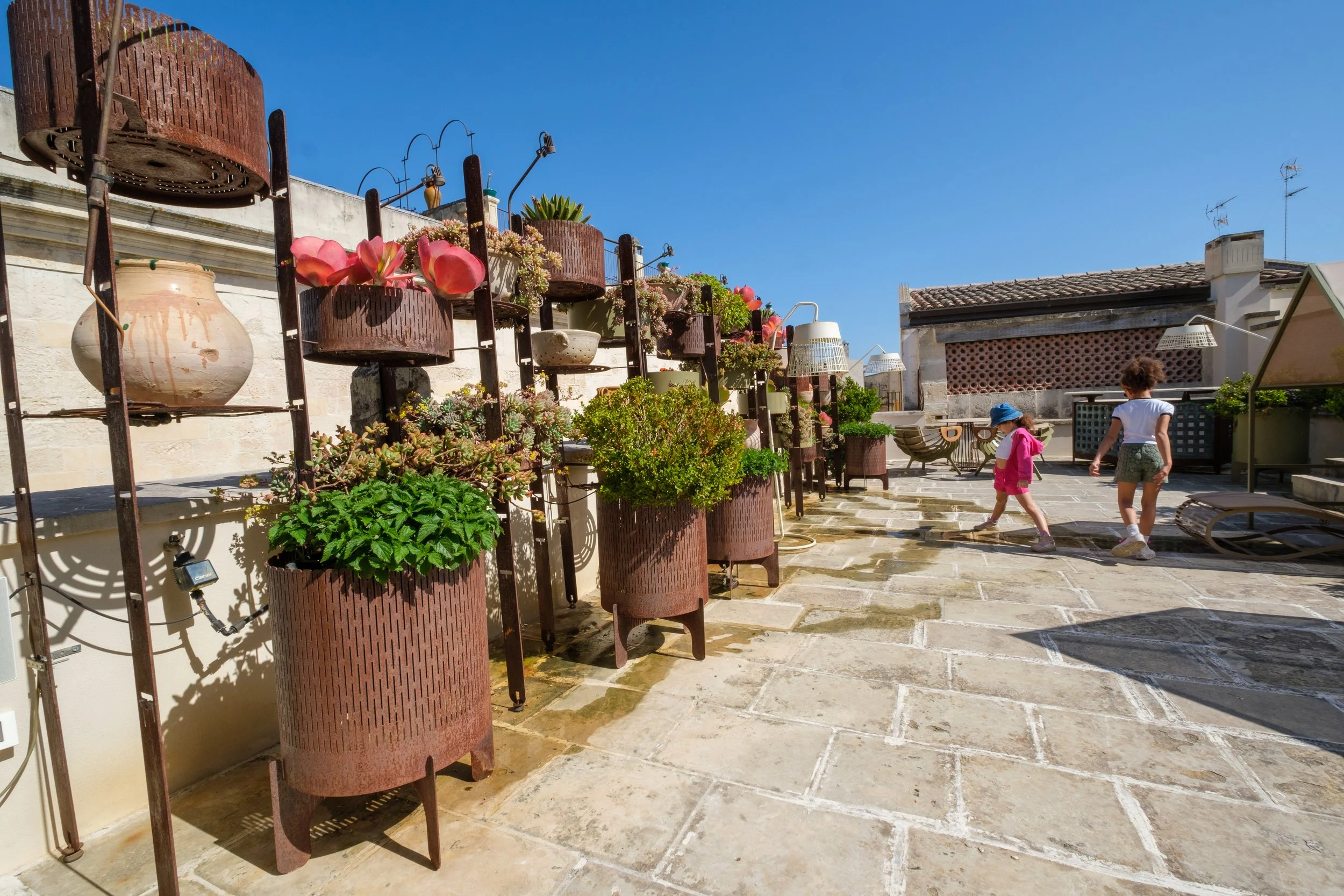 A rooftop terrace with a wall decorated with potted plants and flowers, children playing on the stone floor, and a clear blue sky overhead.
