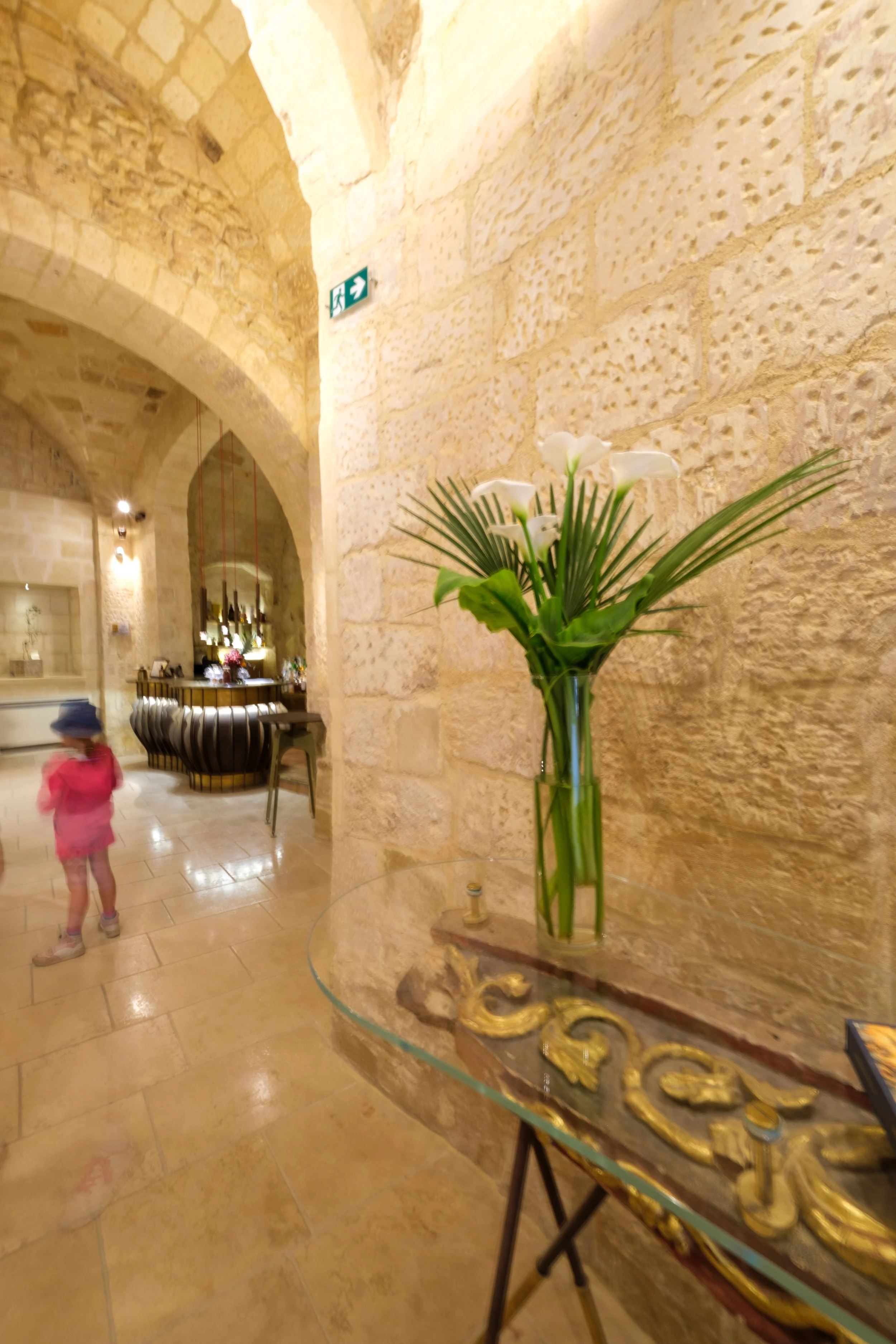 Interior of a building with stone walls, a decorative table with a gold accent, a vase with white flowers and green leaves, and a person wearing a pink jacket and a hat walking by.