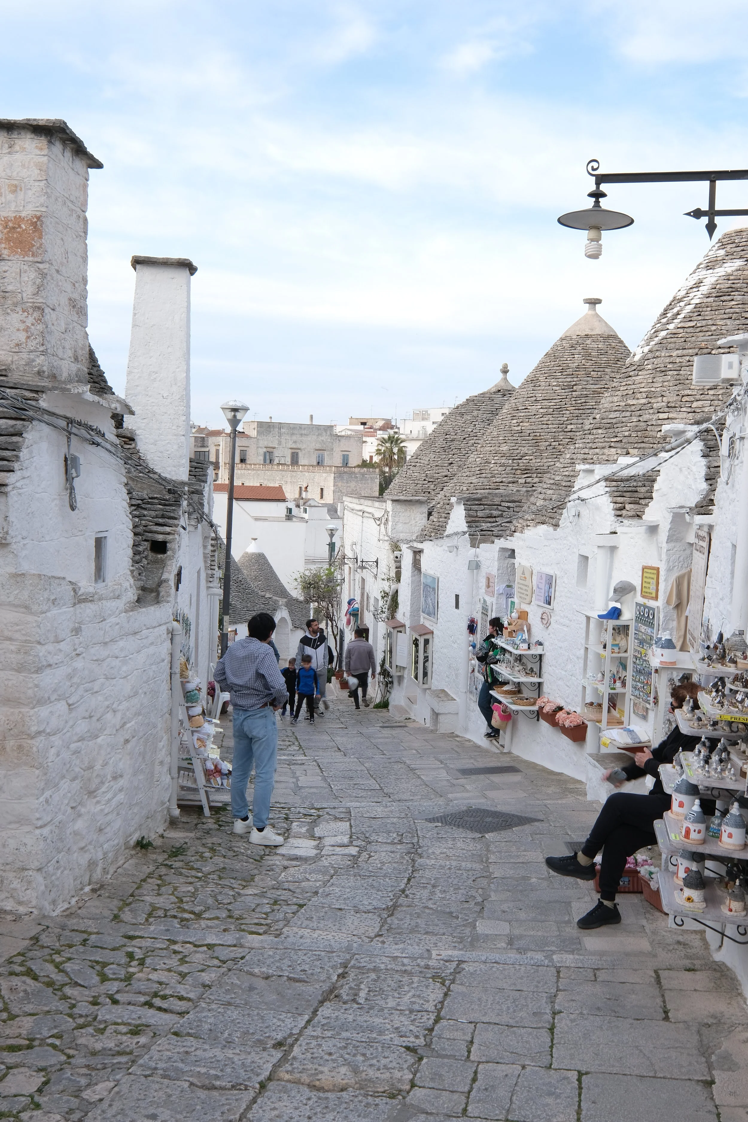 Street scene with white stone buildings and conical roofs, people walking and shopping, outdoor display of souvenirs and crafts, lamp posts, and a cobblestone walkway.
