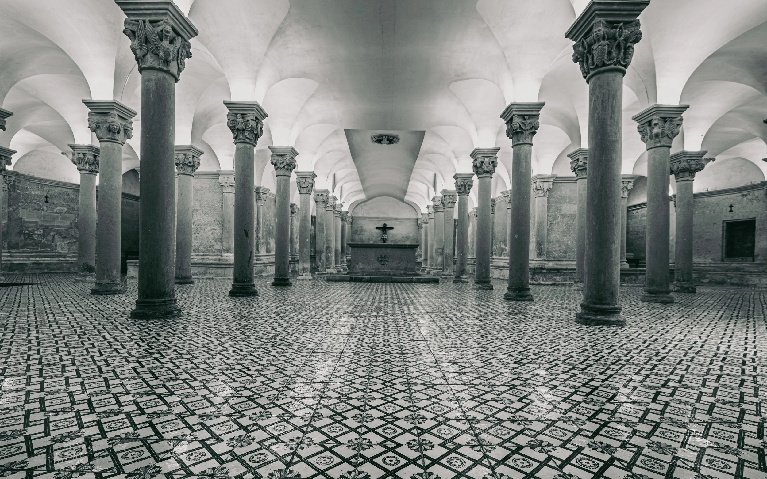 Black and white interior of a historic church with ornate columns, patterned tile floor, and an altar with a crucifix at the rear.