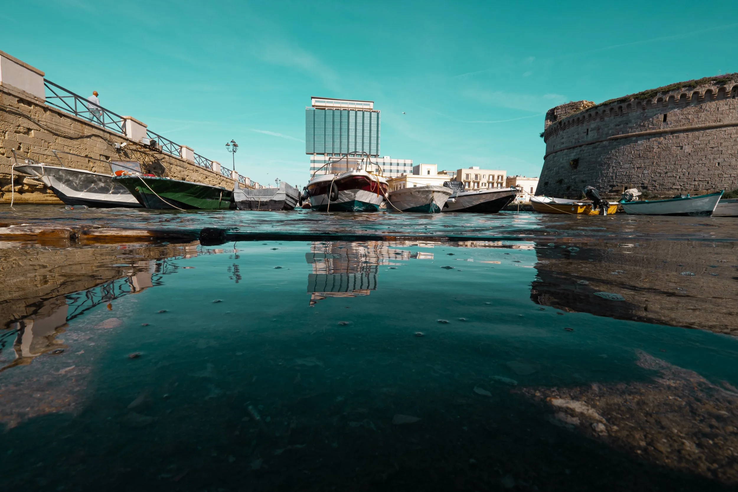 Several small boats docked along a stone waterfront, with a cityscape and blue sky in the background.