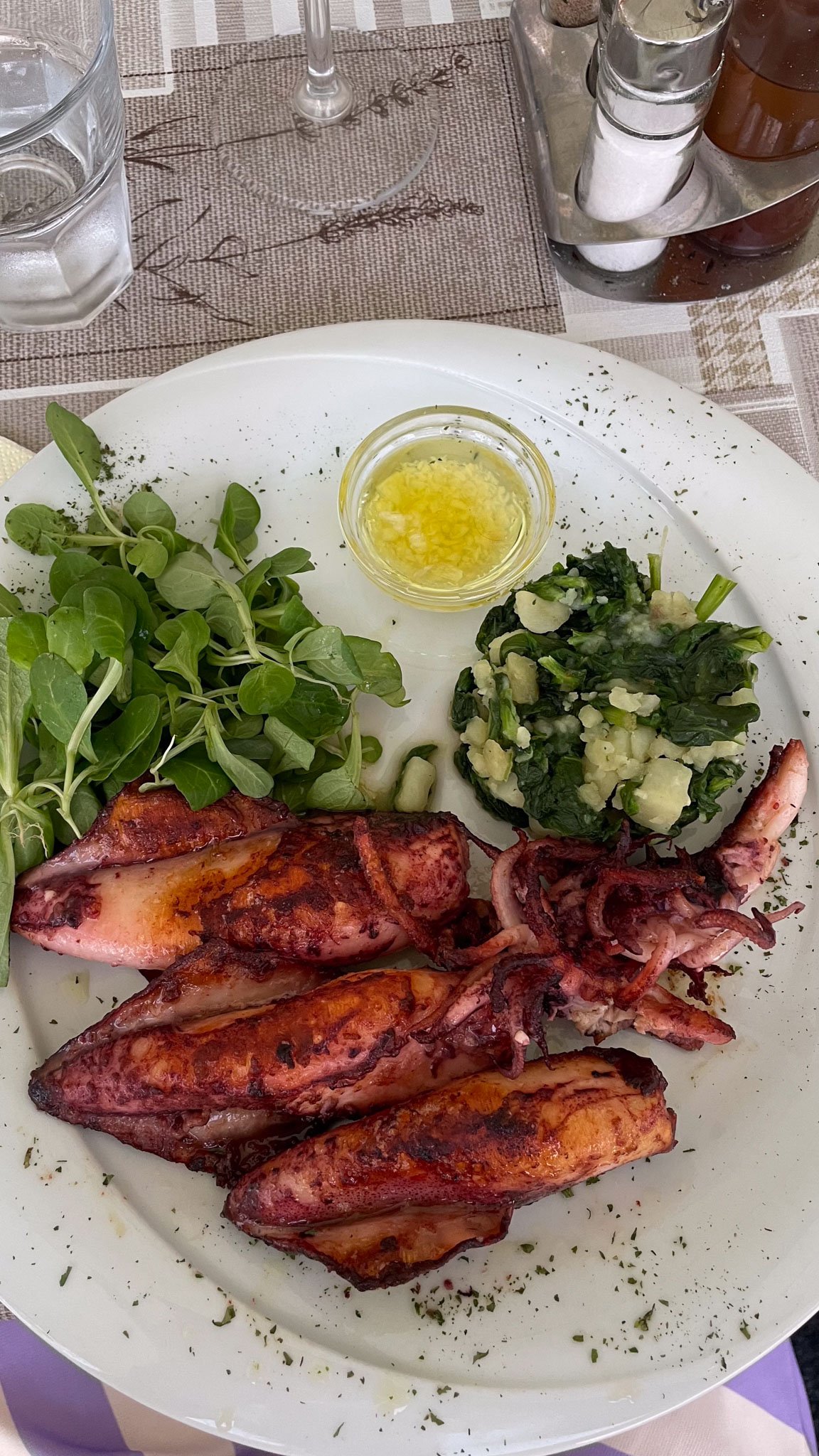 Grilled octopus tentacles served with fresh greens, cooked spinach with cheese, and a small dish of melted butter or oil on a white plate. In the background, there are glasses of water, a wine glass, and condiment shakers on a table set with a woven 