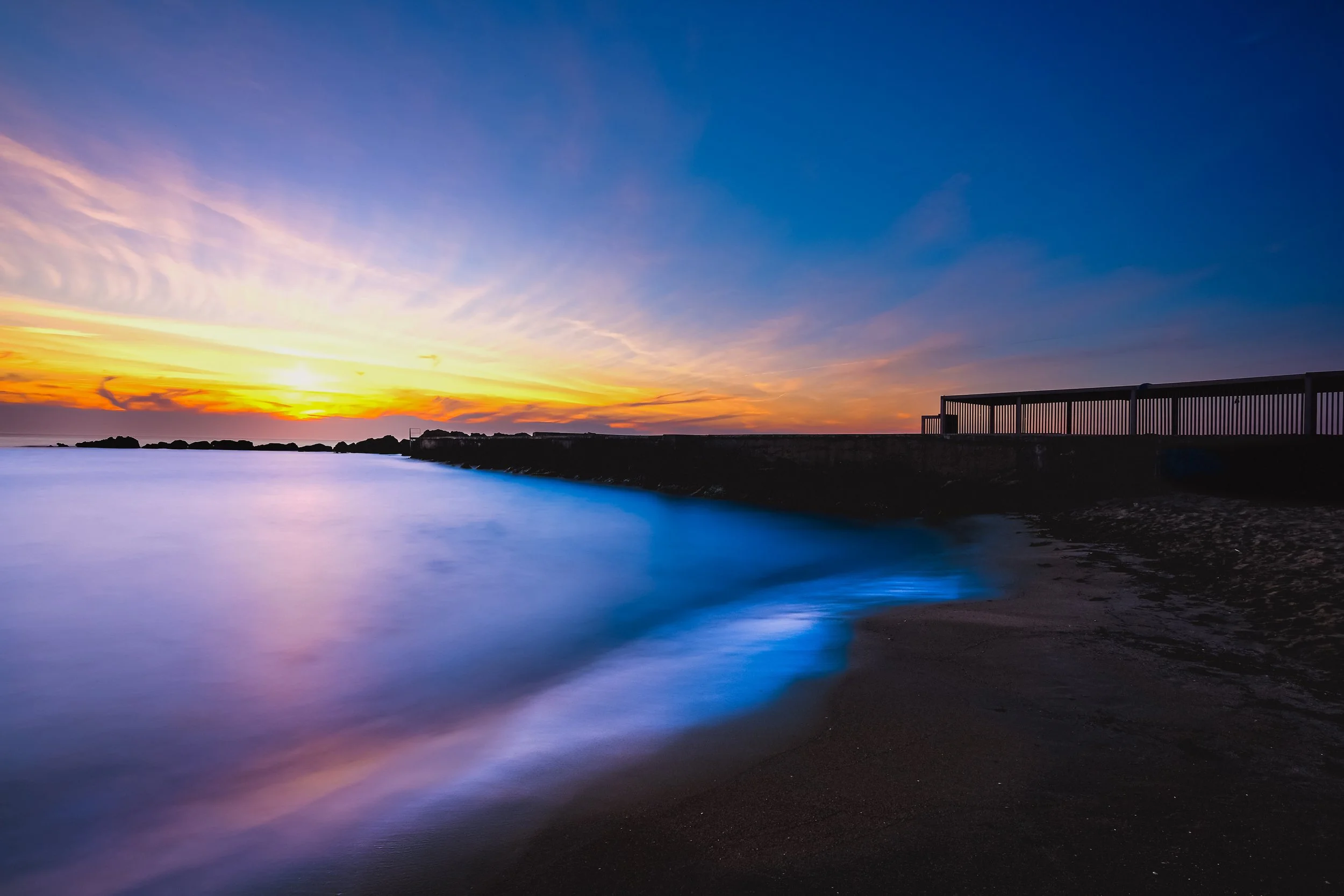A sunset over a body of water with a pier on the right side and rocks in the distance.