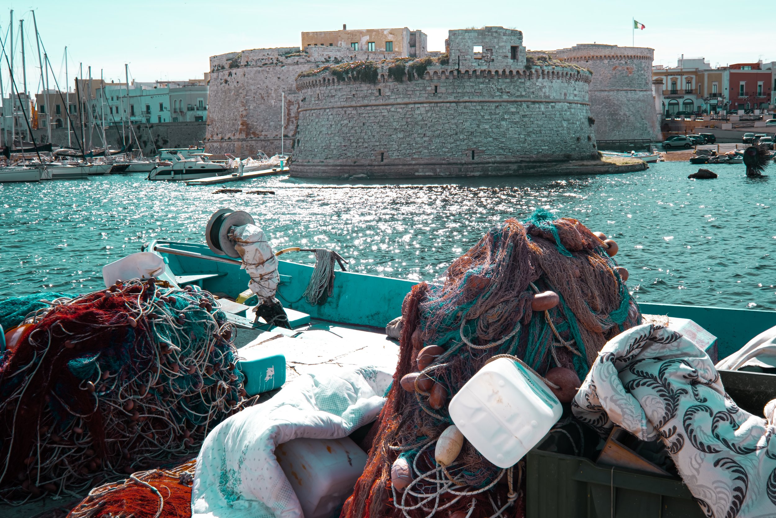 Fishing boats docked near an old stone fortress by the water, with colorful buildings and an Italian flag in the background.