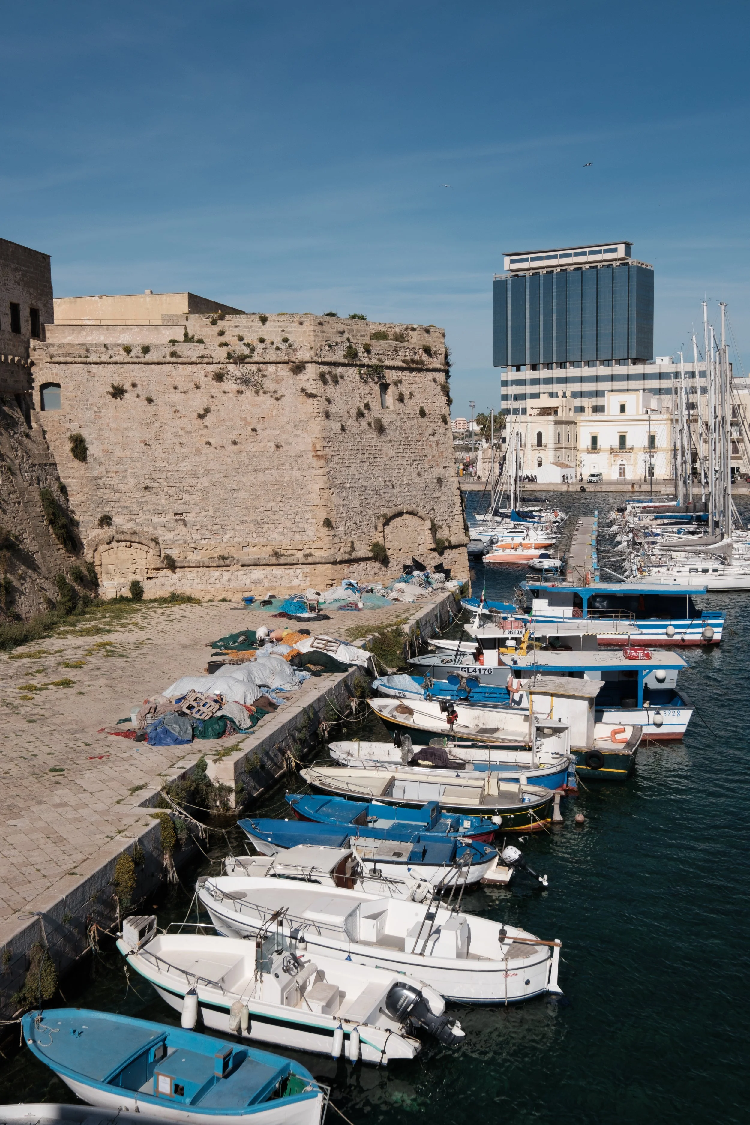 A harbor with several boats docked along the pier, a large stone fortification on the left, a modern glass building in the background, and a clear blue sky with a few birds flying.