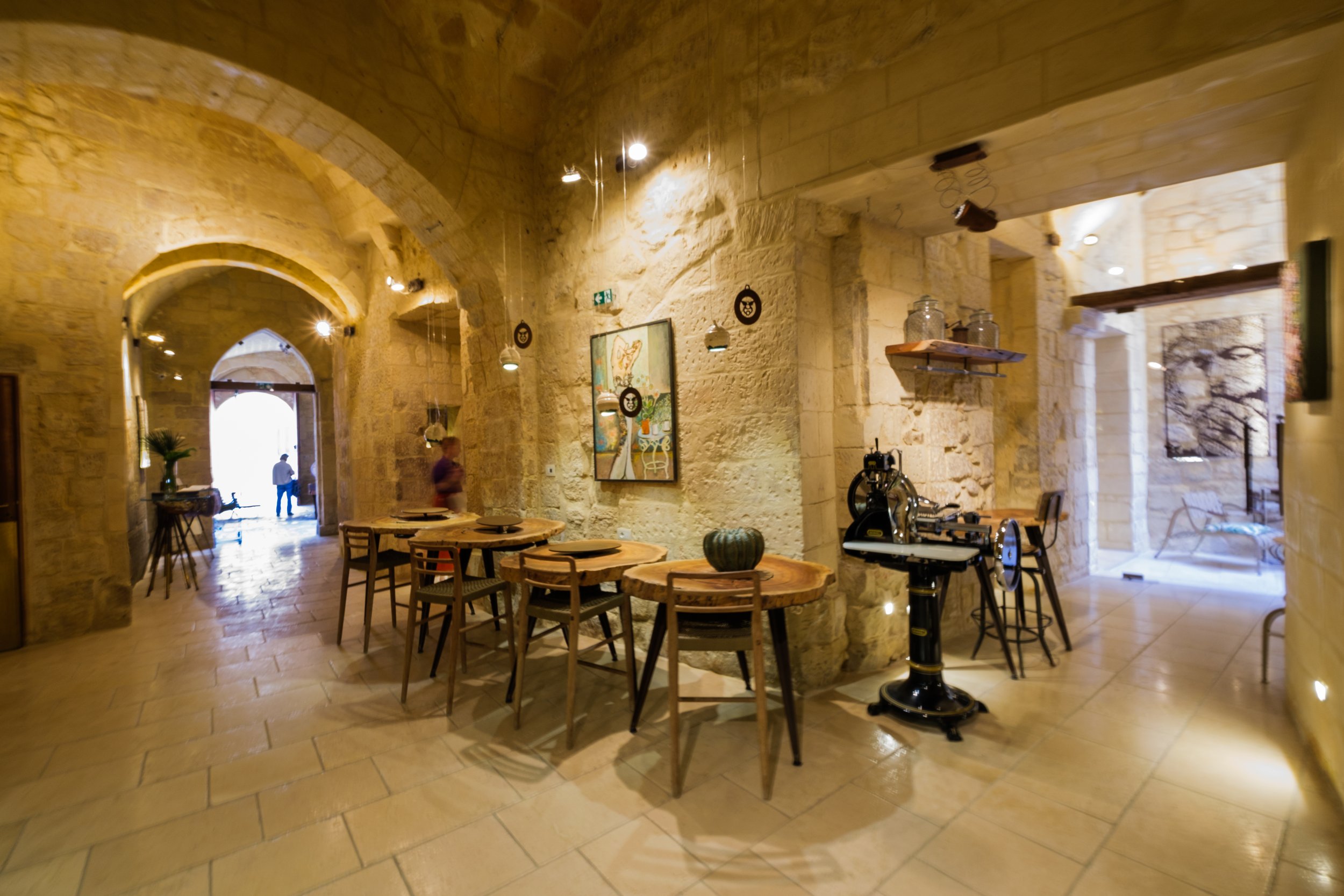 Interior of a rustic stone building with wooden tables and chairs, artwork on the walls, a telescope, and people walking through an arched doorway.