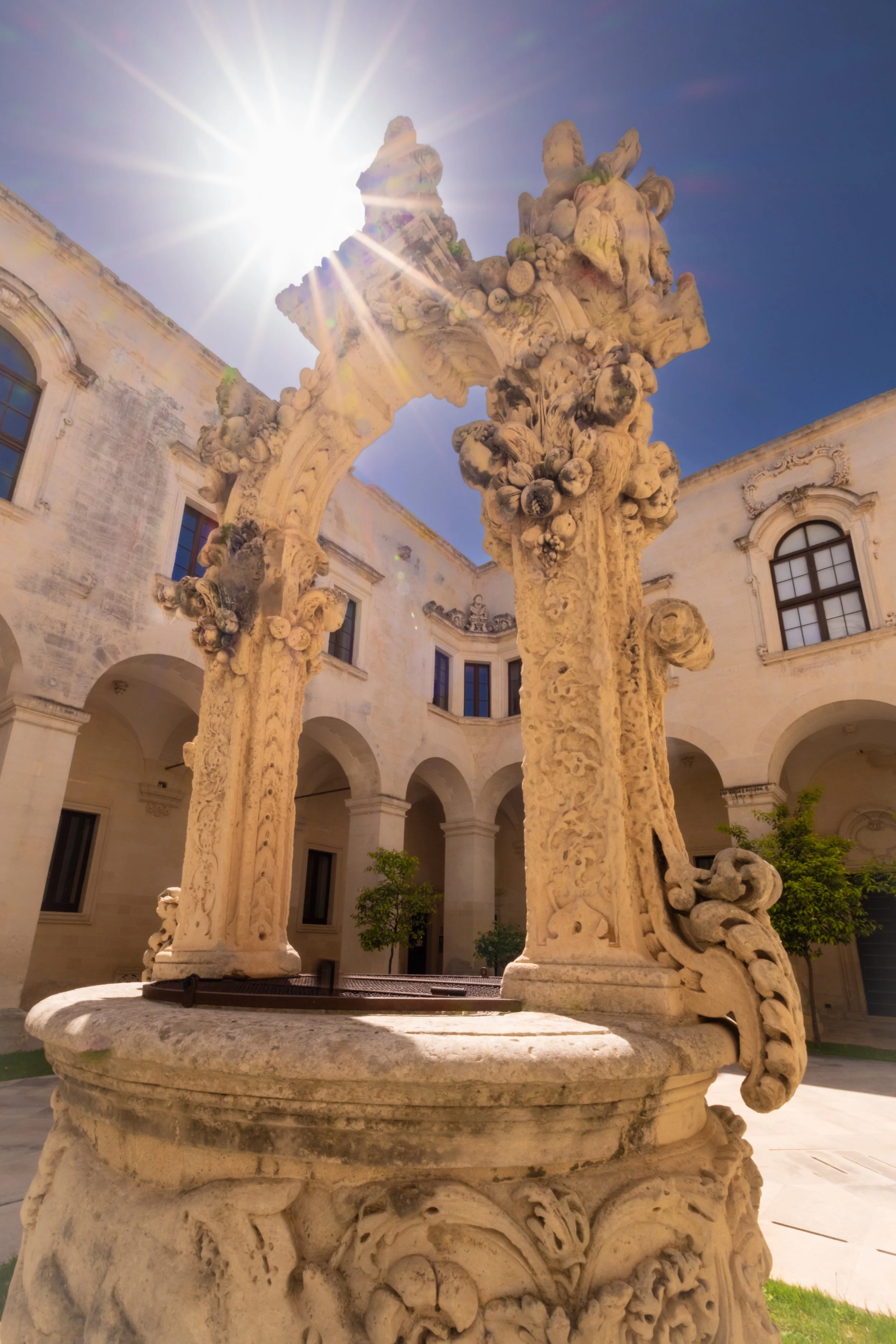 An ornate stone fountain with detailed carvings is shown in a courtyard with a building in the background. The sky is clear and the sun is shining brightly, causing lens flare.