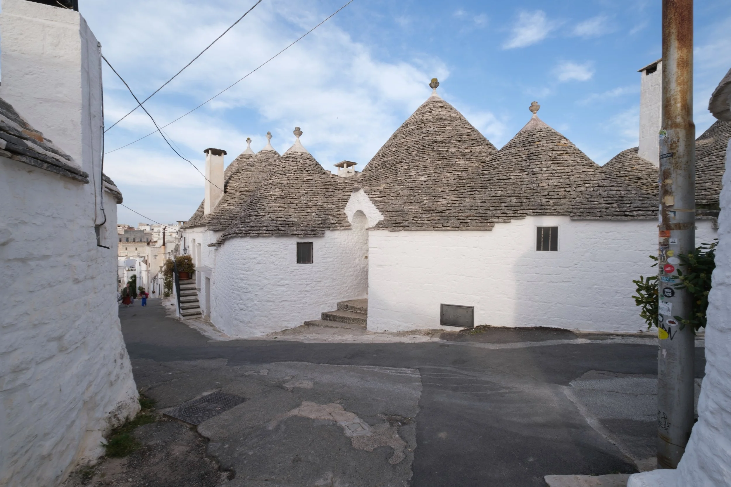 A narrow street in a small town with whitewashed buildings and traditional cone-shaped stone houses with small windows and chimney stacks, under a partly cloudy blue sky.
