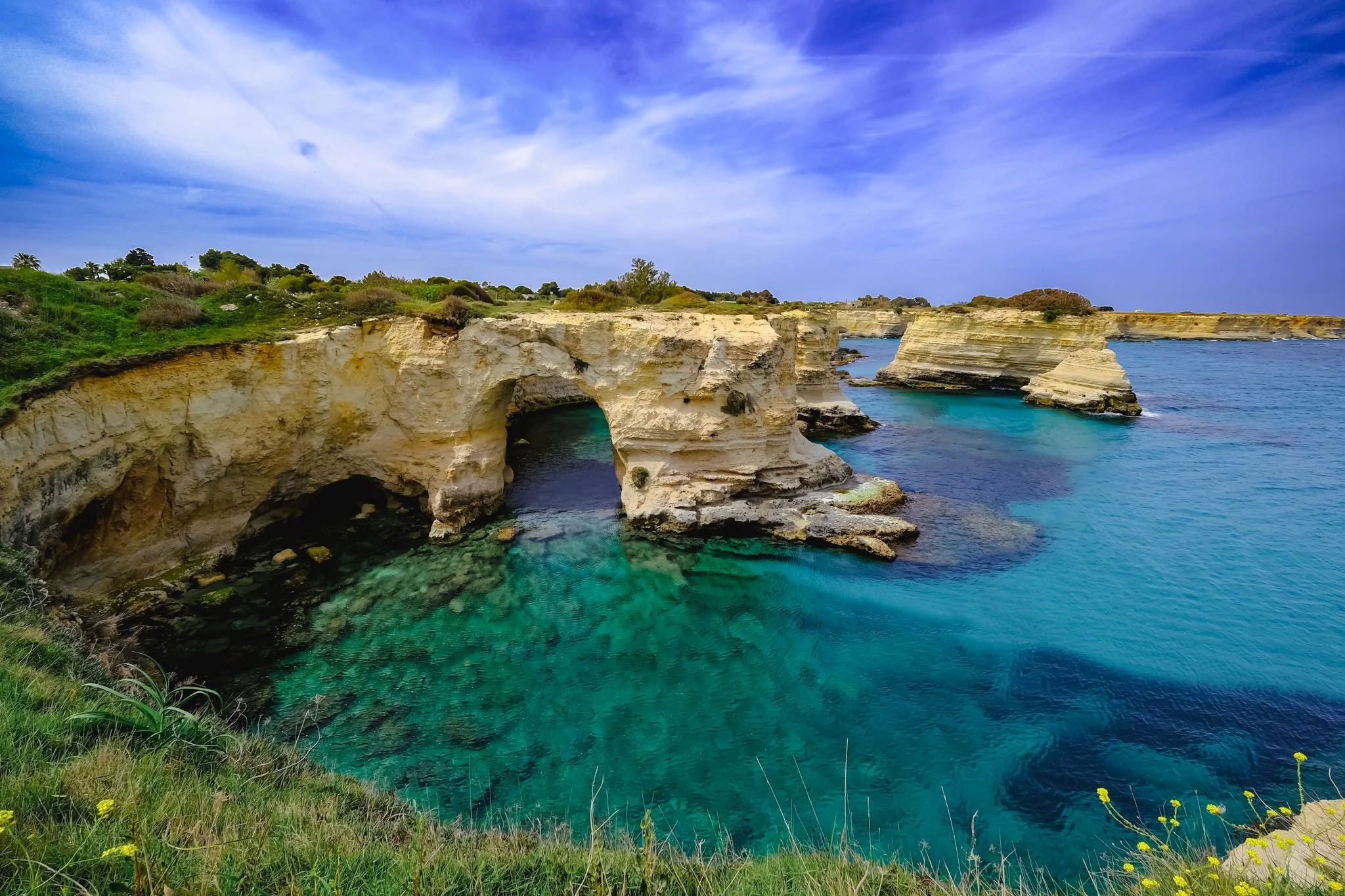 Photo of a coastal landscape with white cliffs, a natural arch formation in the rocks, and turquoise water below. Green vegetation is at the top of the cliffs under a partly cloudy sky.