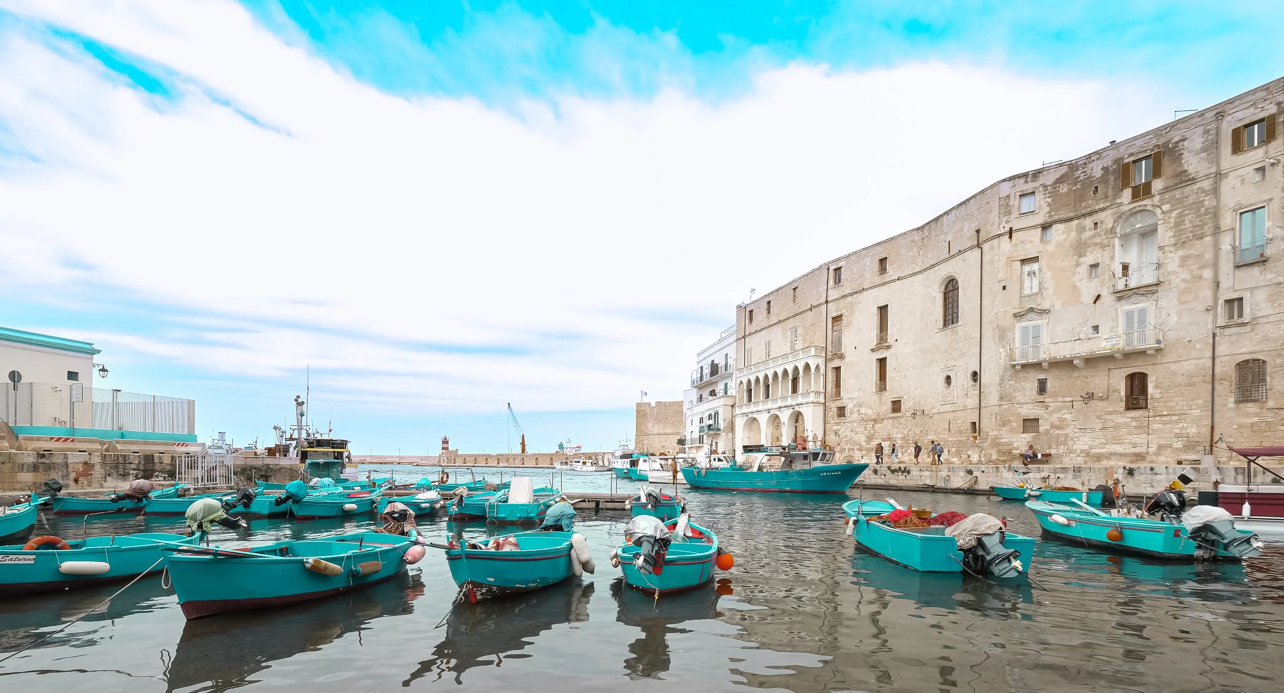 A harbor with small blue boats docked along the water in front of old stone buildings and a clear sky with some clouds.