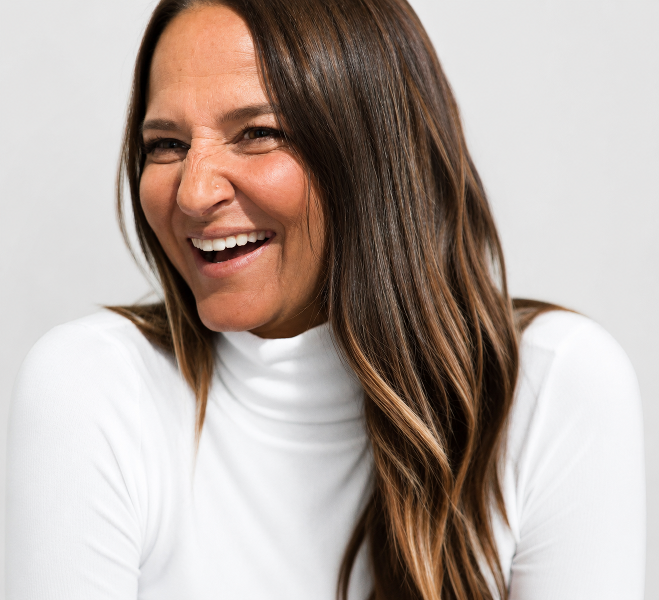 A woman with long brown hair laughing, wearing a white top, against a plain white background.