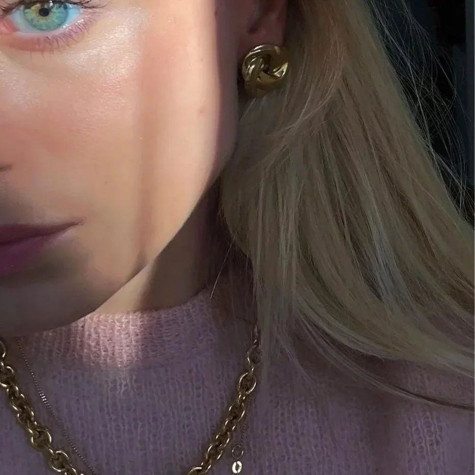 Close-up of a woman's face showing her eye, lips, gold knot earrings, and a gold chain necklace, with a pink sweater and blonde hair.