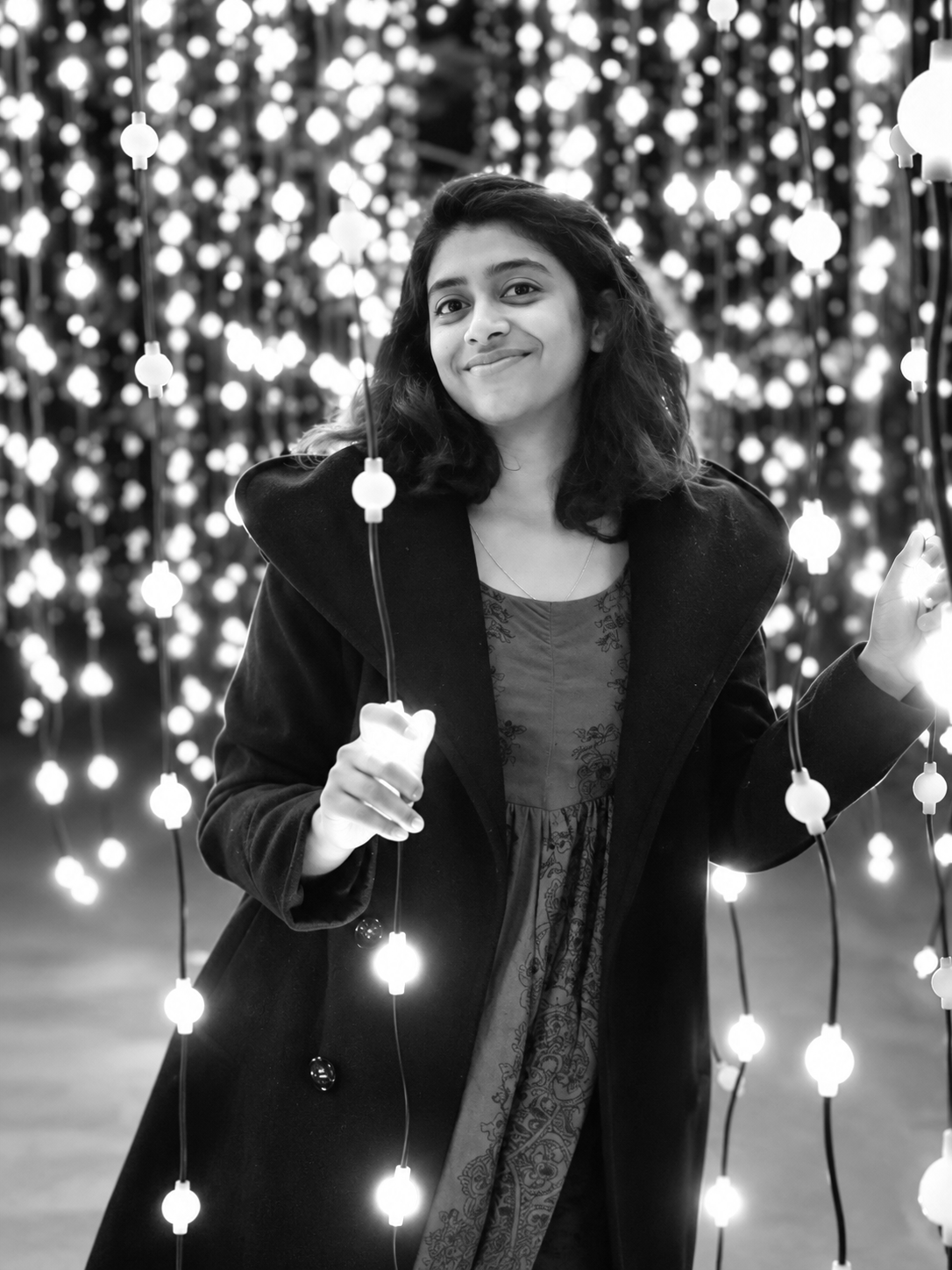 A woman with shoulder-length hair smiling while holding string lights in a dark setting filled with illuminated bulbs.