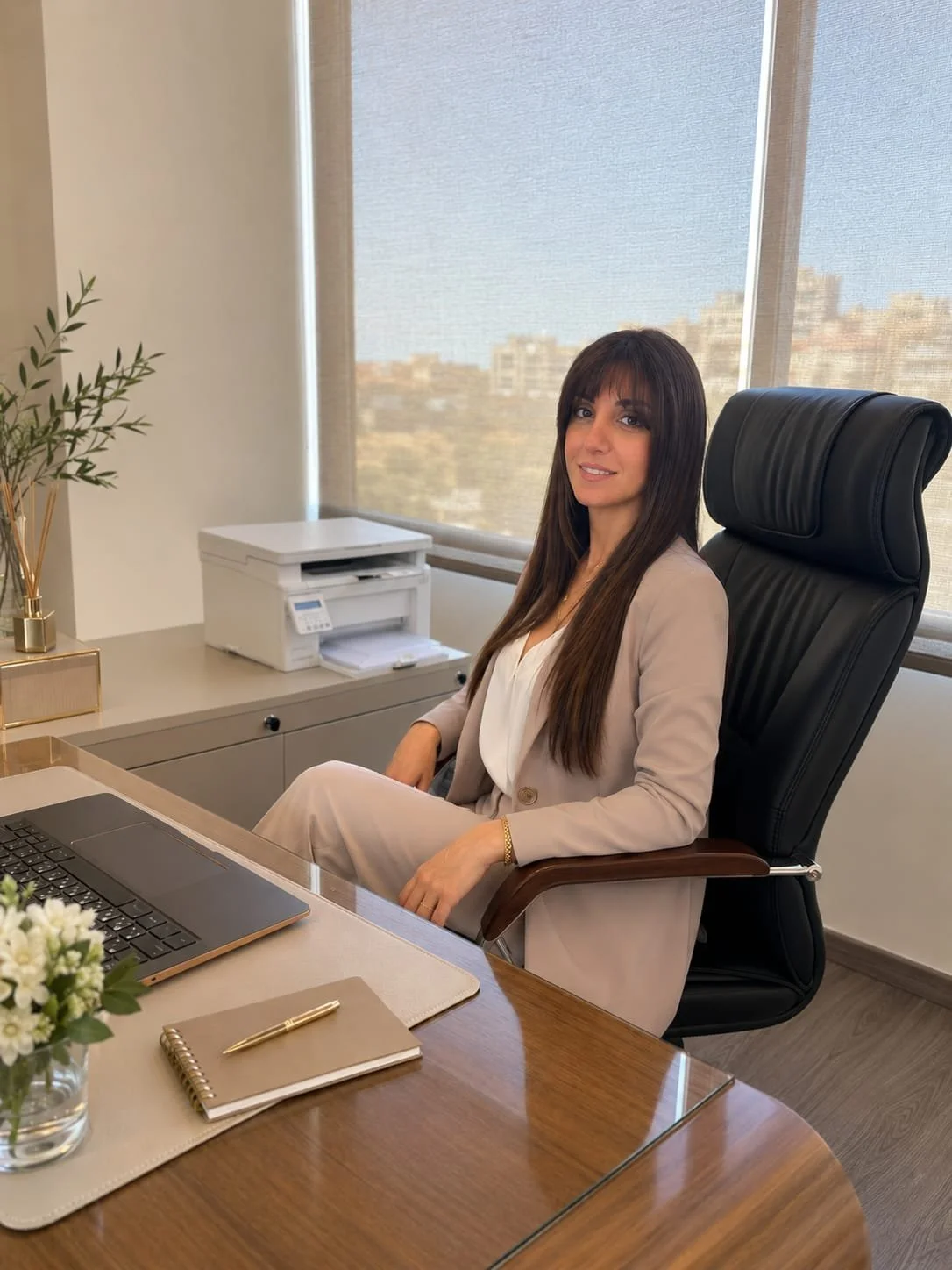 A woman sitting in a black leather office chair at a wooden desk in a modern office, with a window showing a cityscape view behind her, a potted plant, a printer, a laptop, a notepad, and a pen on the desk.