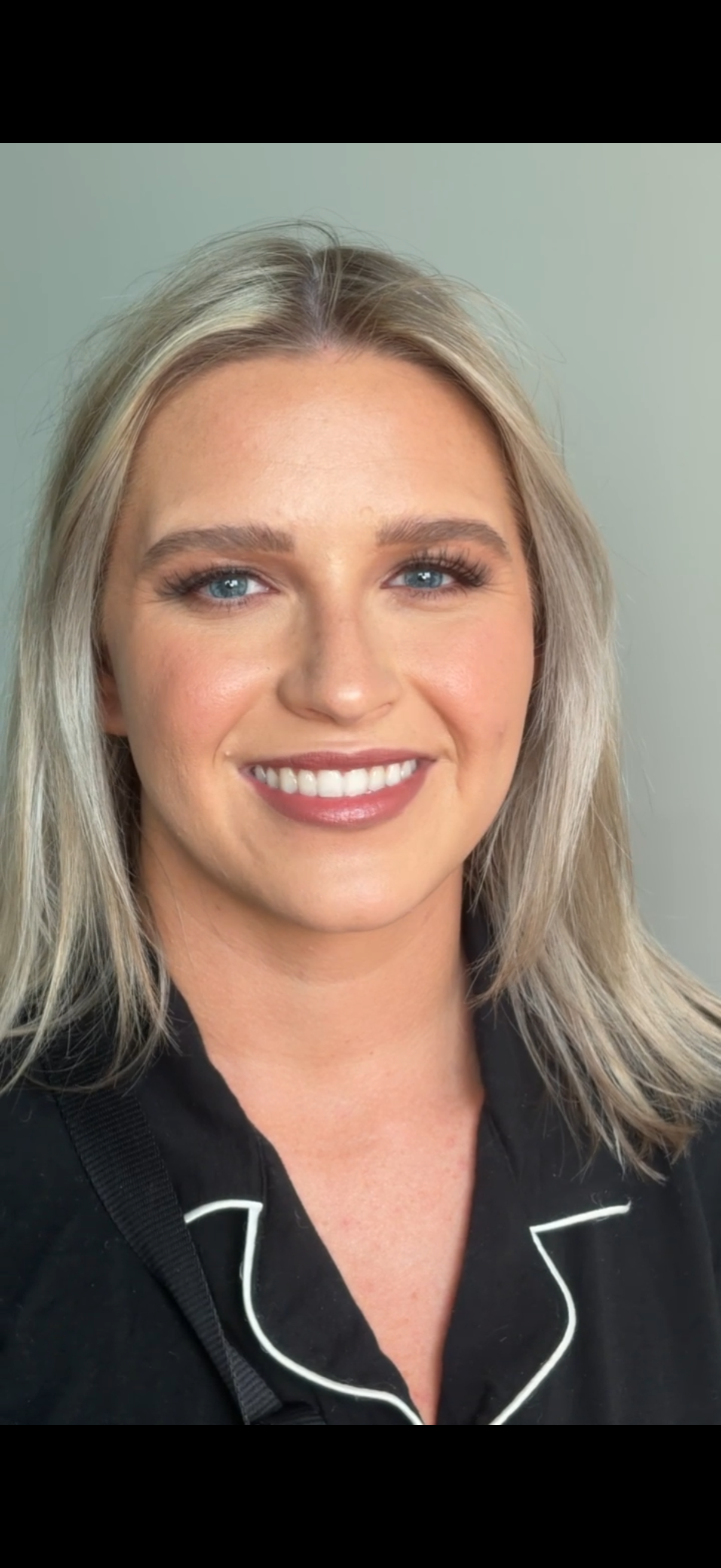 Close-up of a smiling woman with blonde hair, blue eyes, and light skin, wearing a black top with white piping, against a plain background.